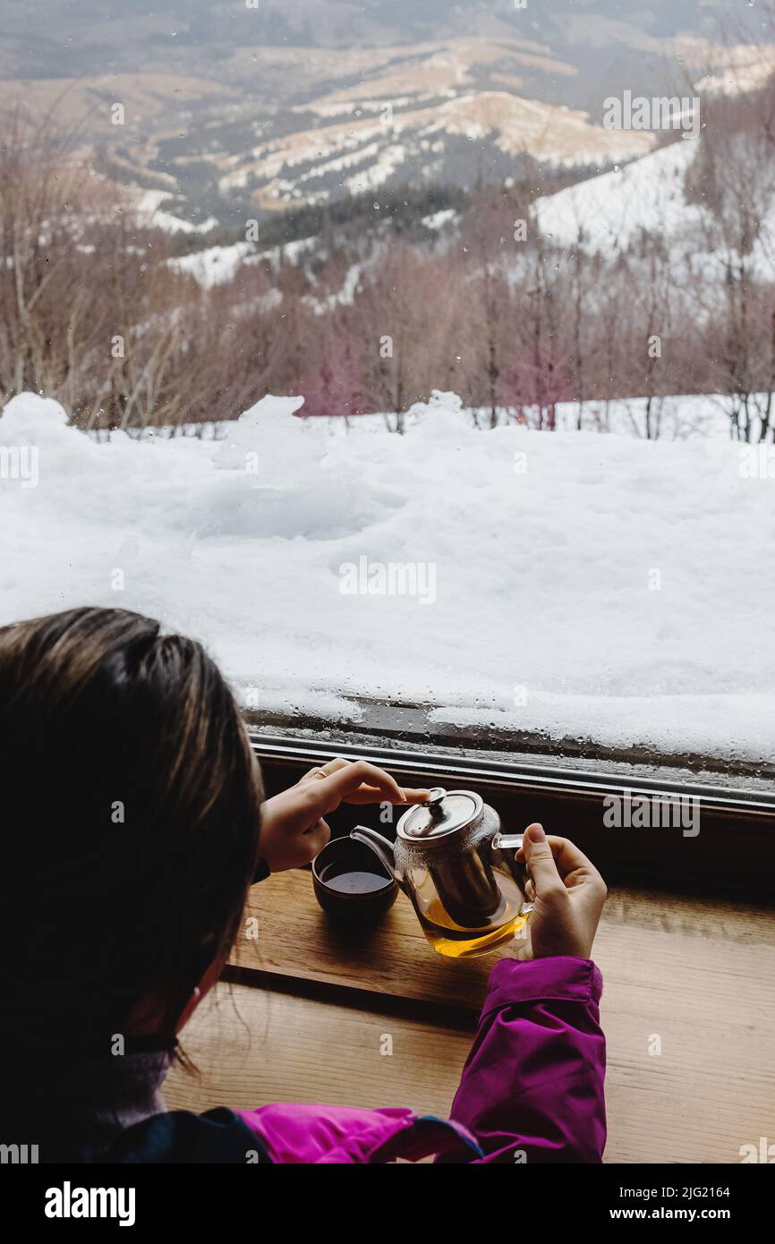 Donna che beve il tè vicino alla grande finestra con vista sulle montagne invernali. Atmosfera accogliente Foto Stock