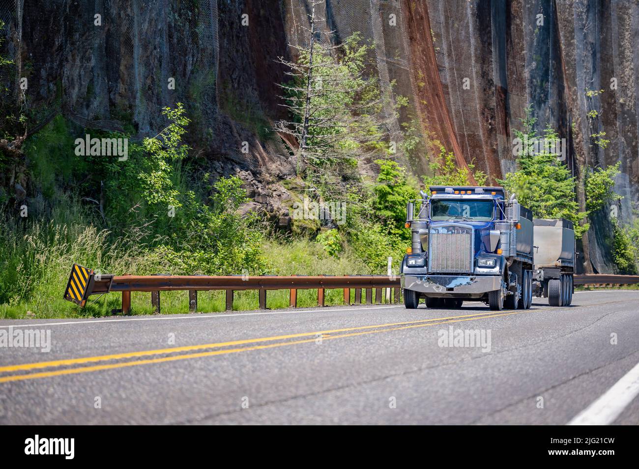 Classic blu American Big RIG Tipper semirimorchio trattore trasporto carico in due punte rimorchi che corrono sulla tortuosa strada di montagna con pareti rocciose e. Foto Stock