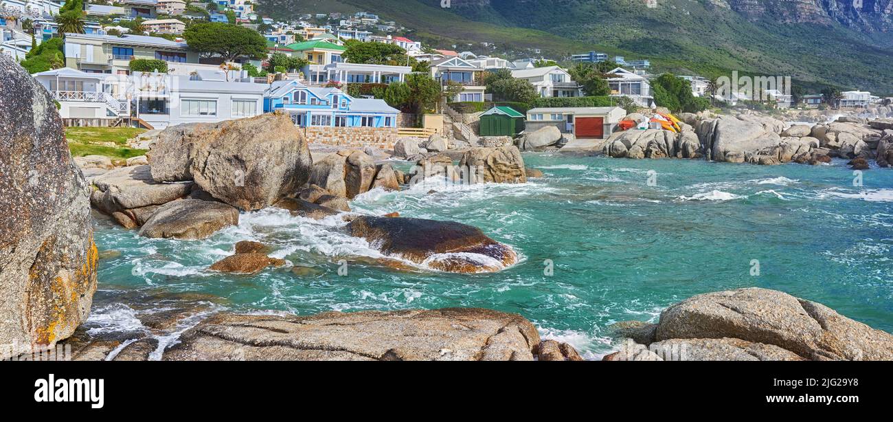 Vista panoramica del mare, rocce ed edifici residenziali a Camps Bay Beach, Città del Capo, Sud Africa. Onde dell'oceano marea che si lavano sopra le rocce di litorale e. Foto Stock