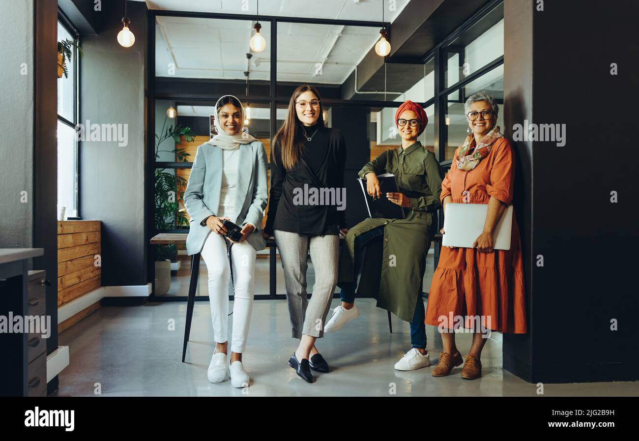 Designer multiculturali sorridenti alla fotocamera in una sala riunioni d'ufficio. Imprenditrici creative che lavorano in squadra in un ambiente di lavoro inclusivo. Gruppo di hap Foto Stock