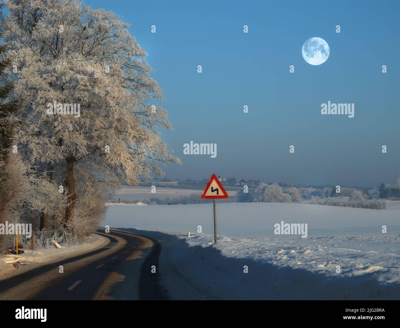 La strada tortuosa su un paesaggio rurale invernale con un cartello sul lato della strada vuota per la sicurezza. Una strada scivolosa e bagnata circondata da neve su un Foto Stock