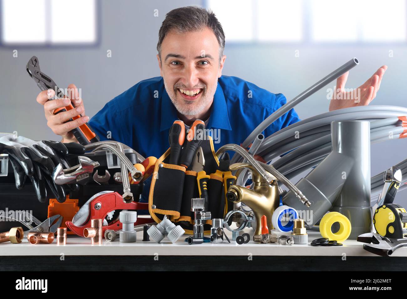 Esempio di materiali e utensili idraulici sul banco di lavoro con idraulico sorridente dietro in magazzino industriale. Vista frontale. Composizione orizzontale. Foto Stock