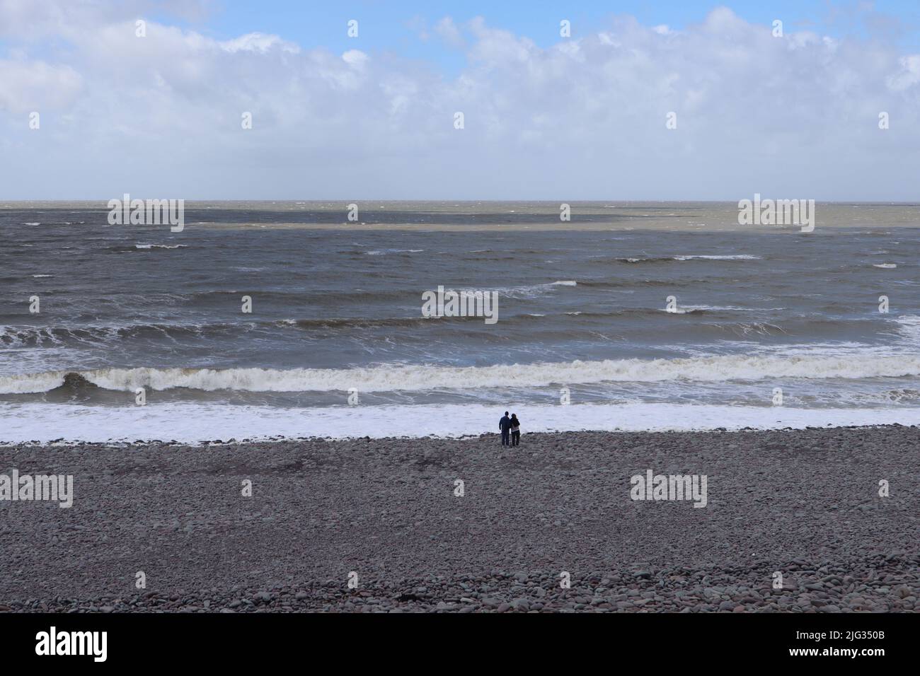 In lontananza, una coppia si trova sul bordo delle acque e guarda le onde e il mare a Bossington Beach in Somerset, Inghilterra Foto Stock