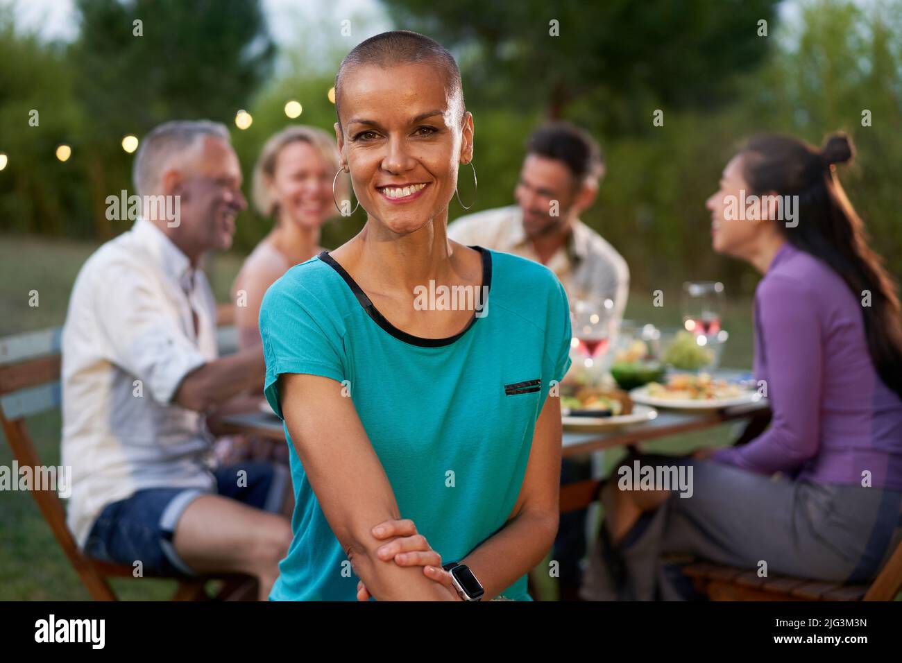 Ritratto di una donna con testa rasata che guarda la fotocamera. Gruppo di amici che cenano all'aperto su un ristorante con terrazza o casa sul cortile. Amici che hanno Foto Stock