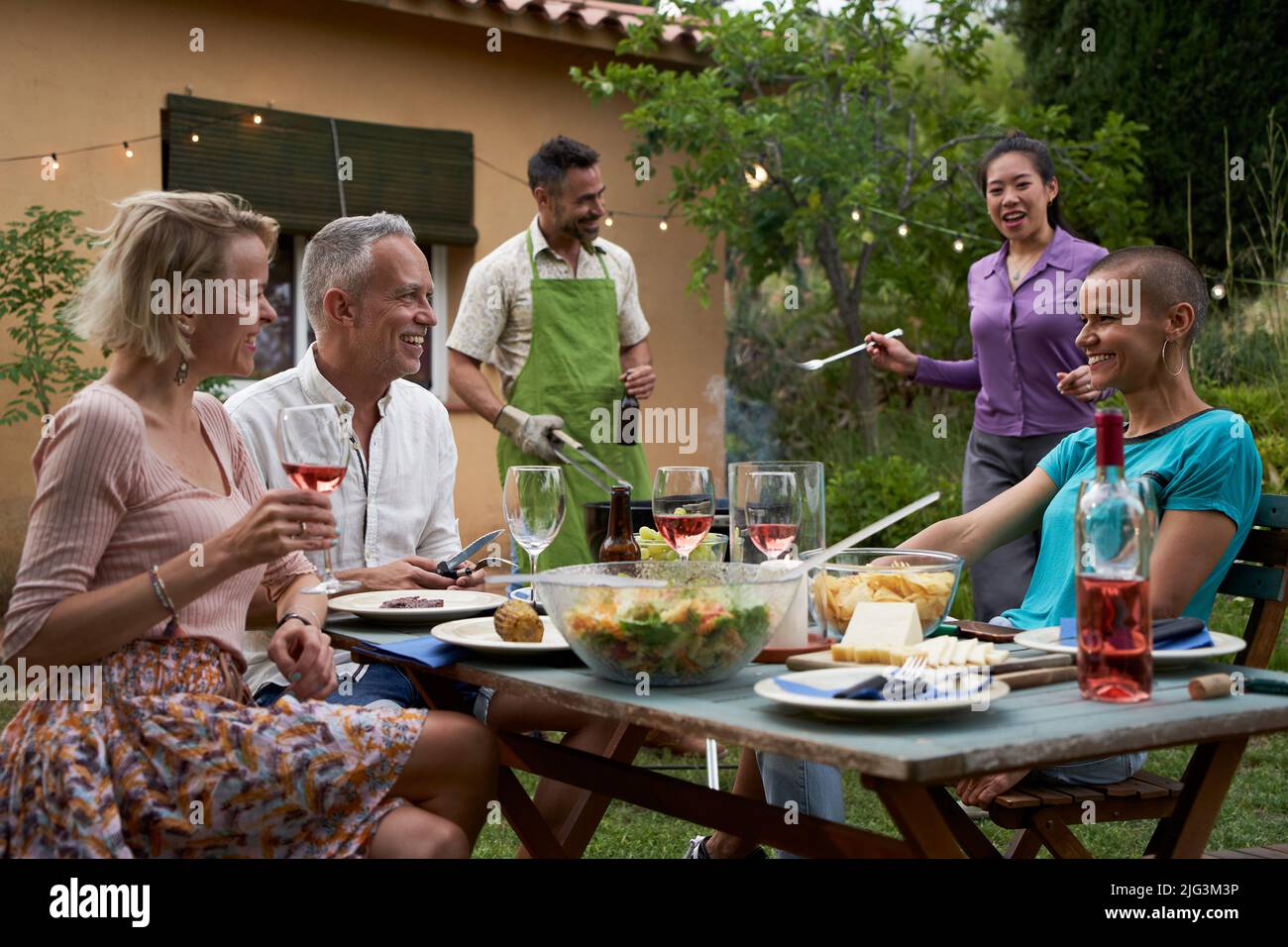 Gente di mezza età che cena e divertimento bevendo il vino rosso insieme nel giardino di casa. Amici felici che mangiano cibo barbecue nel cortile. Stile di vita di amicizia Foto Stock