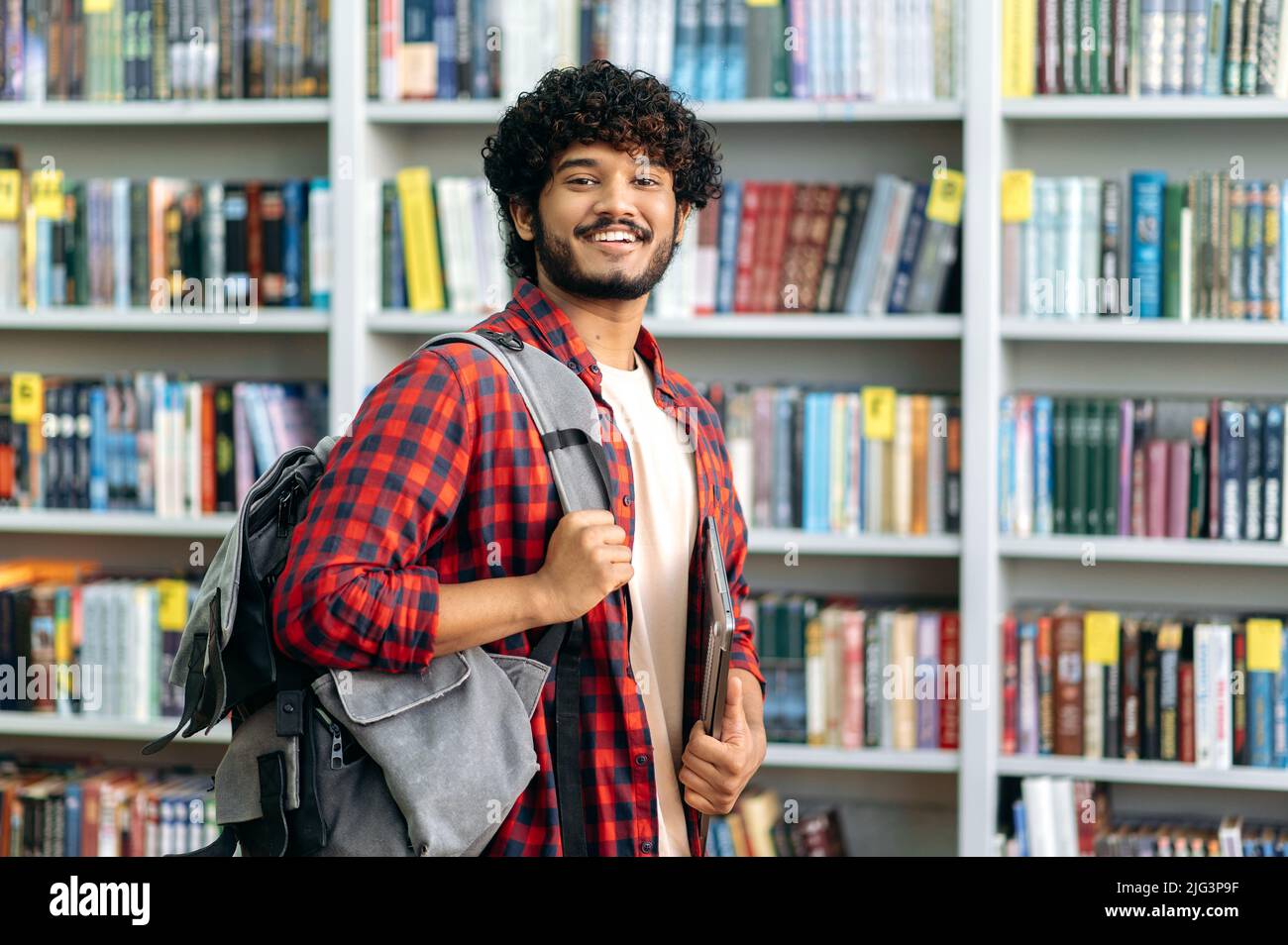 Bello orgoglioso riccio capelli indiano o arabo maschio studente di università, in abbigliamento casual, con zaino e laptop, si erge in una biblioteca sullo sfondo di scaffali, guarda la macchina fotografica, sorride Foto Stock