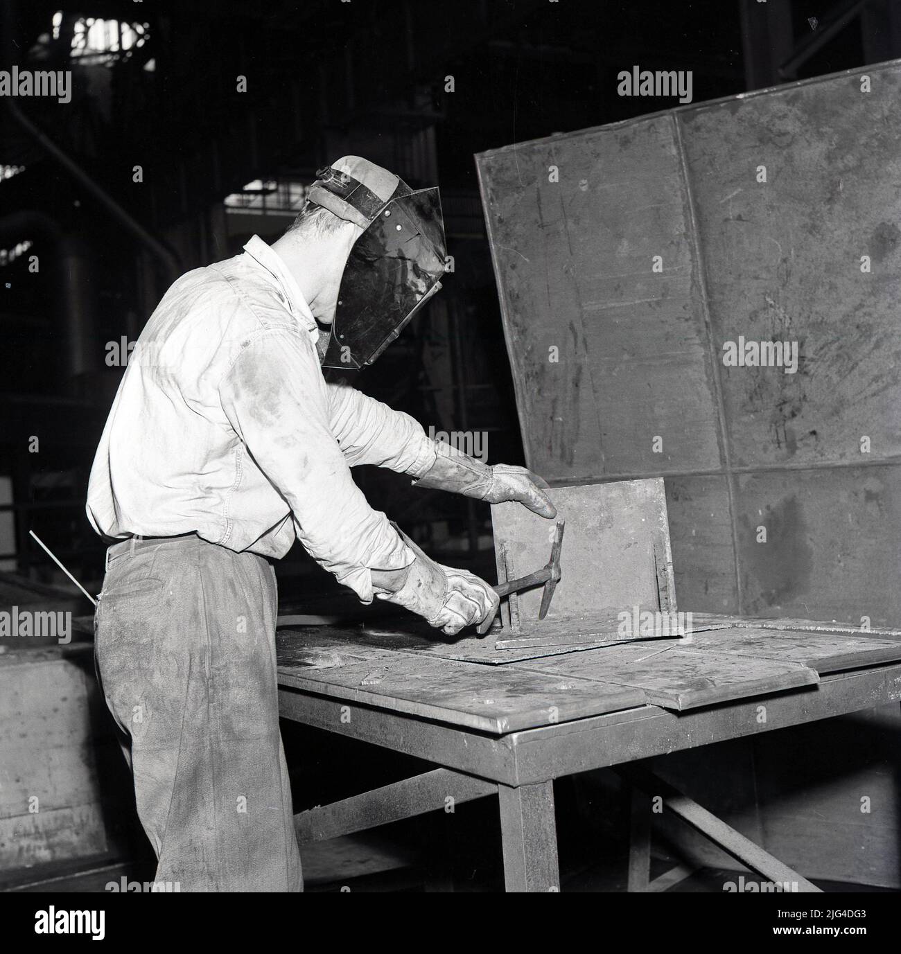 1950s, storico, operaio in acciaio maschio con visiera protettiva che lavora su un banco. Foto Stock