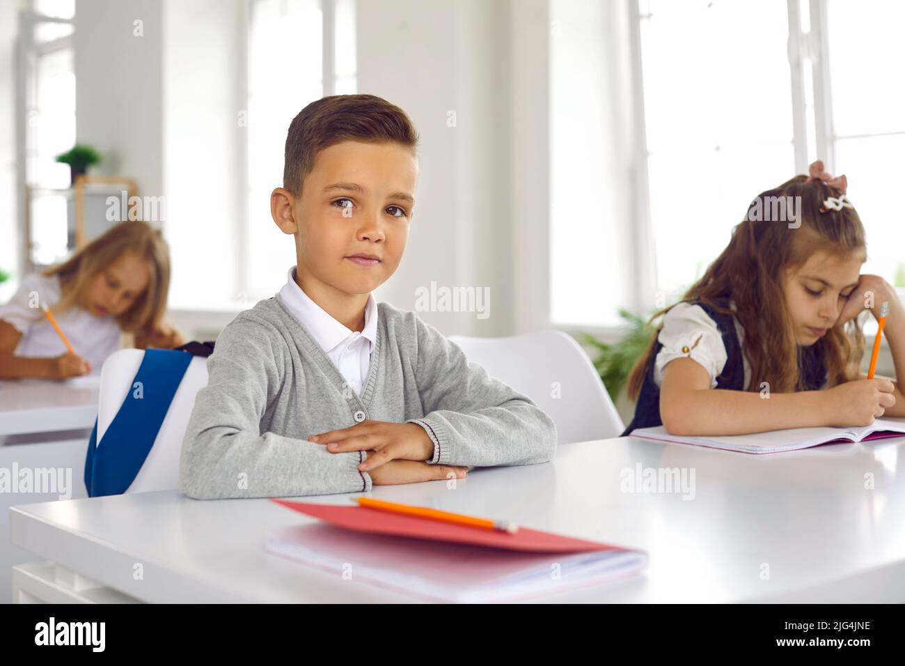 Ritratto di carino sorridente scolaro seduto in aula con le braccia ripiegate in piano sulla scrivania. Foto Stock