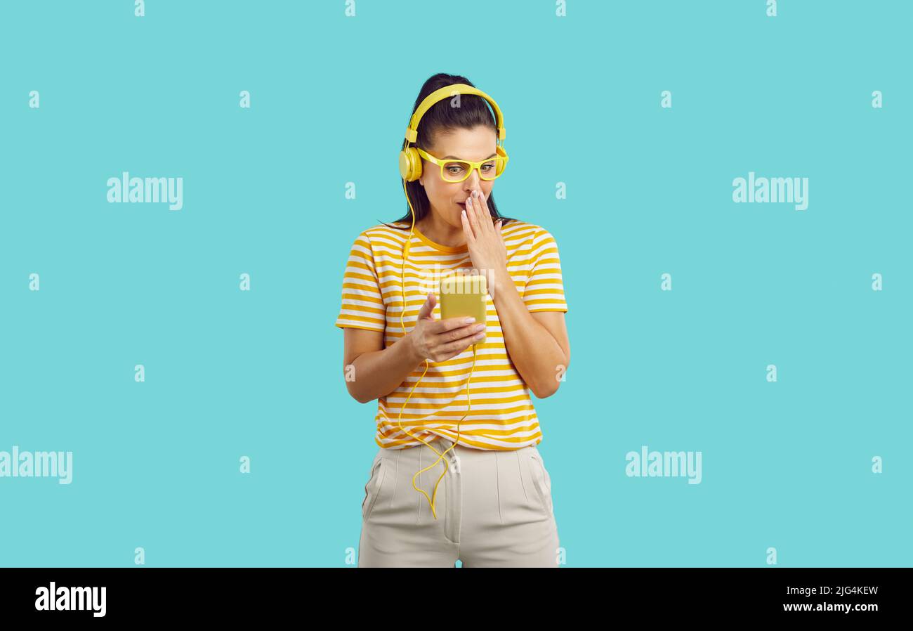Giovane bella donna con cuffie che guardano qualcosa di incredibile sul telefono copre la bocca in shock, sembra timido, in piedi su sfondo blu. Foto Stock