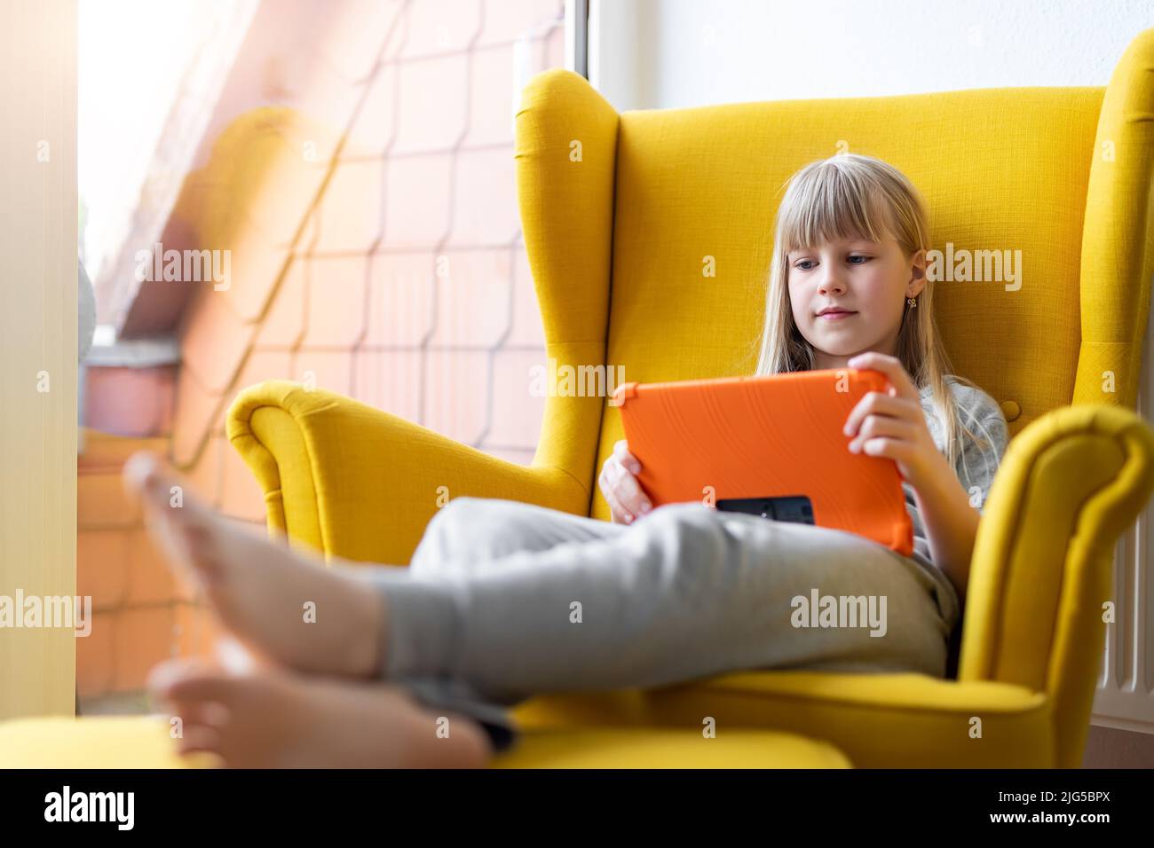 Ritratto di simpatica bionda ragazza piccola goda seduto in morbida poltrona gialla giocare a gioco o guardare film TV show utilizzando tablet Foto Stock