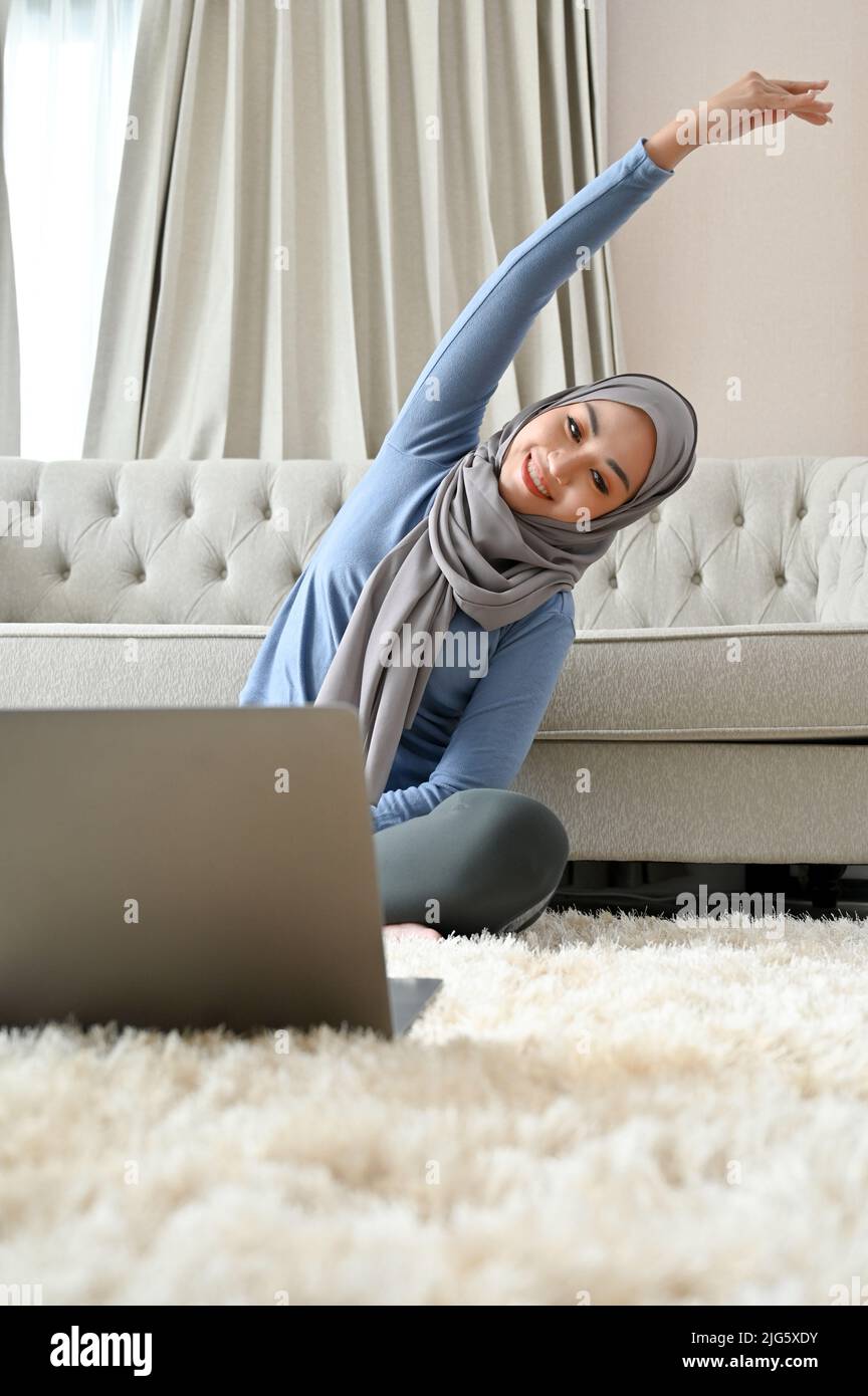 Bella donna musulmana asiatica millenaria che indossa hijab, braccio allungante, yoga imparante pone sul Internet attraverso il suo computer portatile nel vivo r Foto Stock