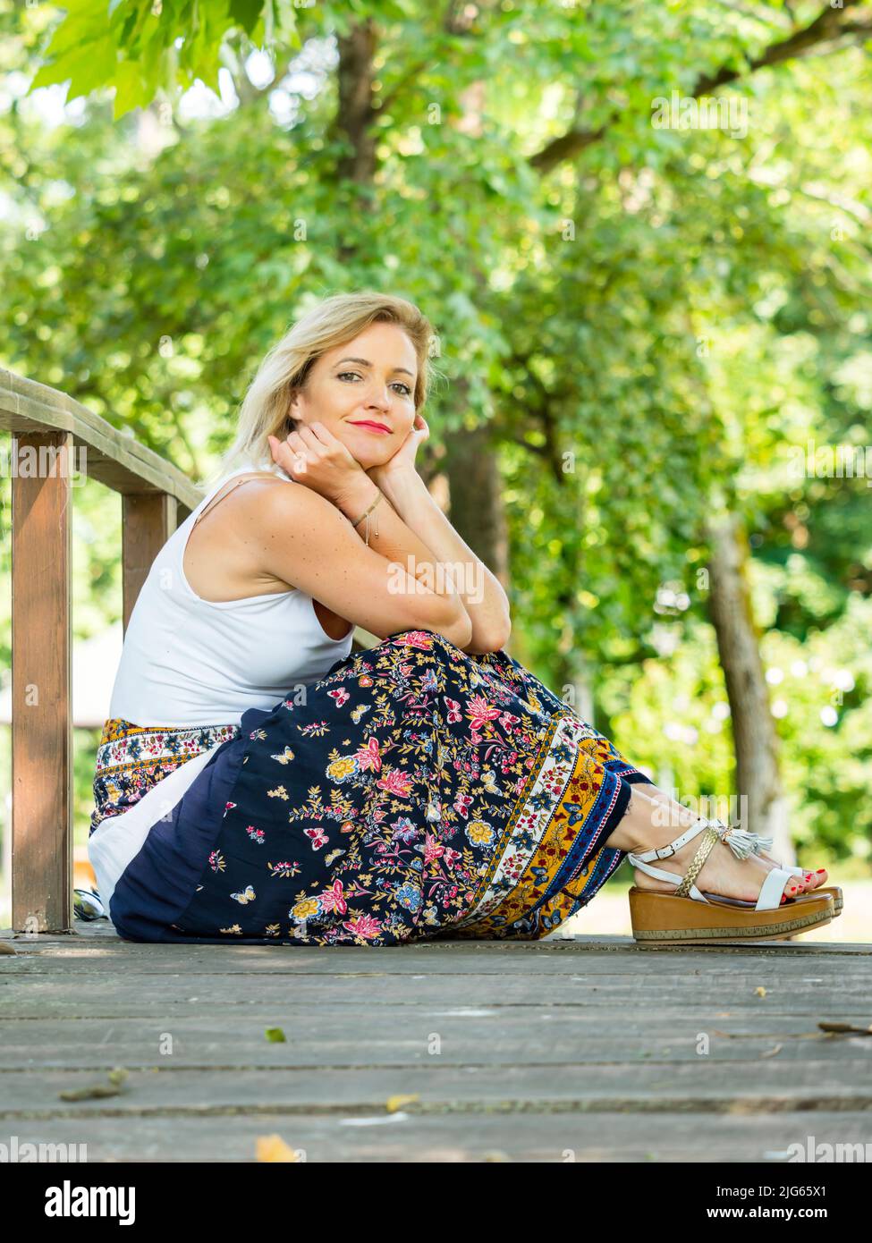Capelli biondi donna 30s è seduta seduta seduta su piede-ponte di legno passerella guardando la macchina fotografica lieve sorriso Foto Stock