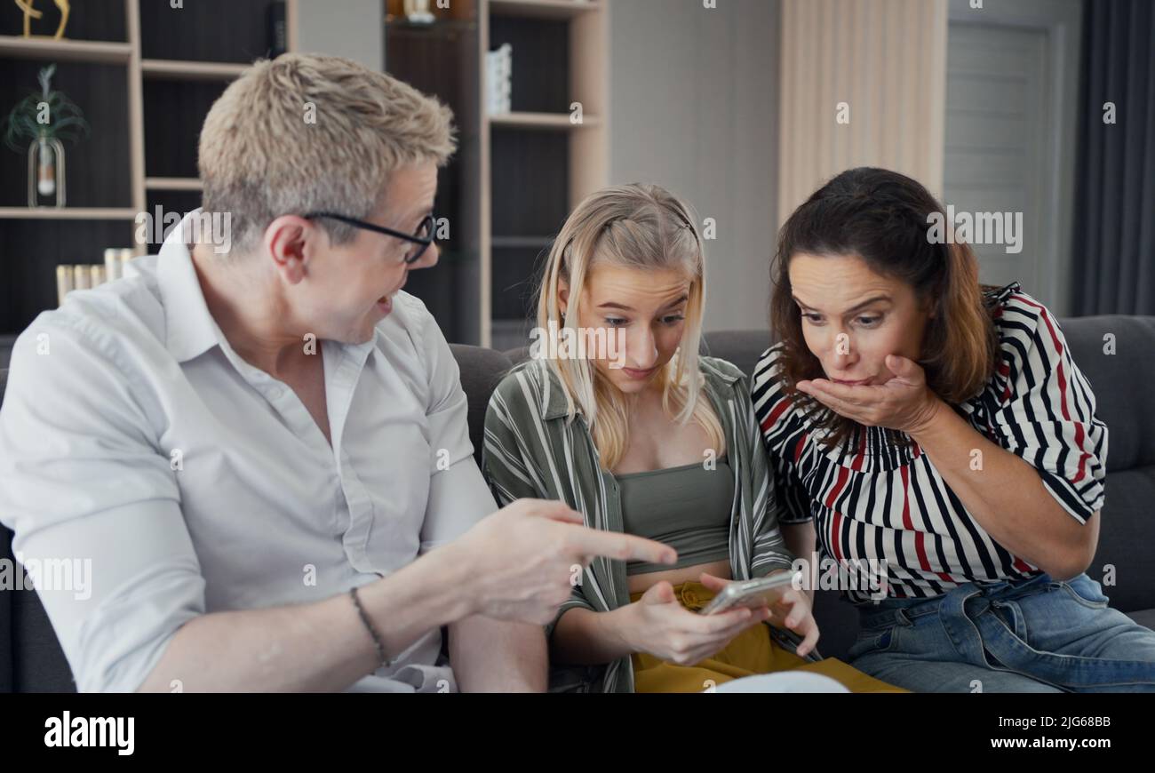 I genitori caucasici seduti sul divano e la figlia eccitata usando lo smart phone, sono felici e si chiedono in qualcosa sullo schermo con il volto strano in livi Foto Stock