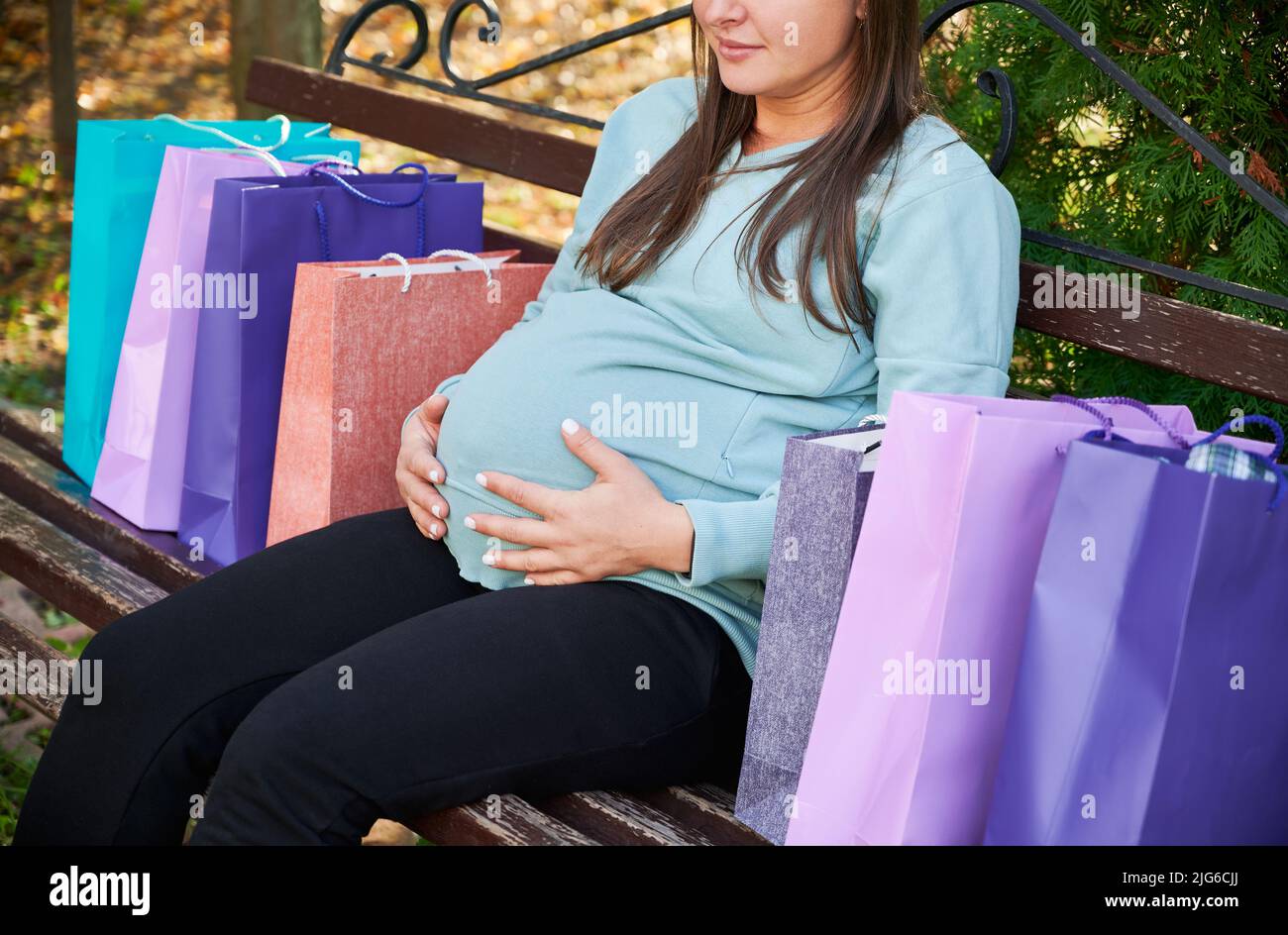 Donna incinta che si rilassa su panca di legno, mettendo le mani sul suo ventre e ponendo borse regalo con acquisti su entrambi i lati. Concetto di periodo meraviglioso di gravidanza nella vita della donna. Vista ritagliata. Foto Stock