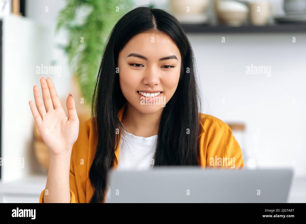 Positivo felice bella cinese giovane donna, seduta a un tavolo con un portatile a casa in cucina, parlare in una videoconferenza online, ondeggiando, chiacchierando con colleghi o amici, sorride amichevole Foto Stock