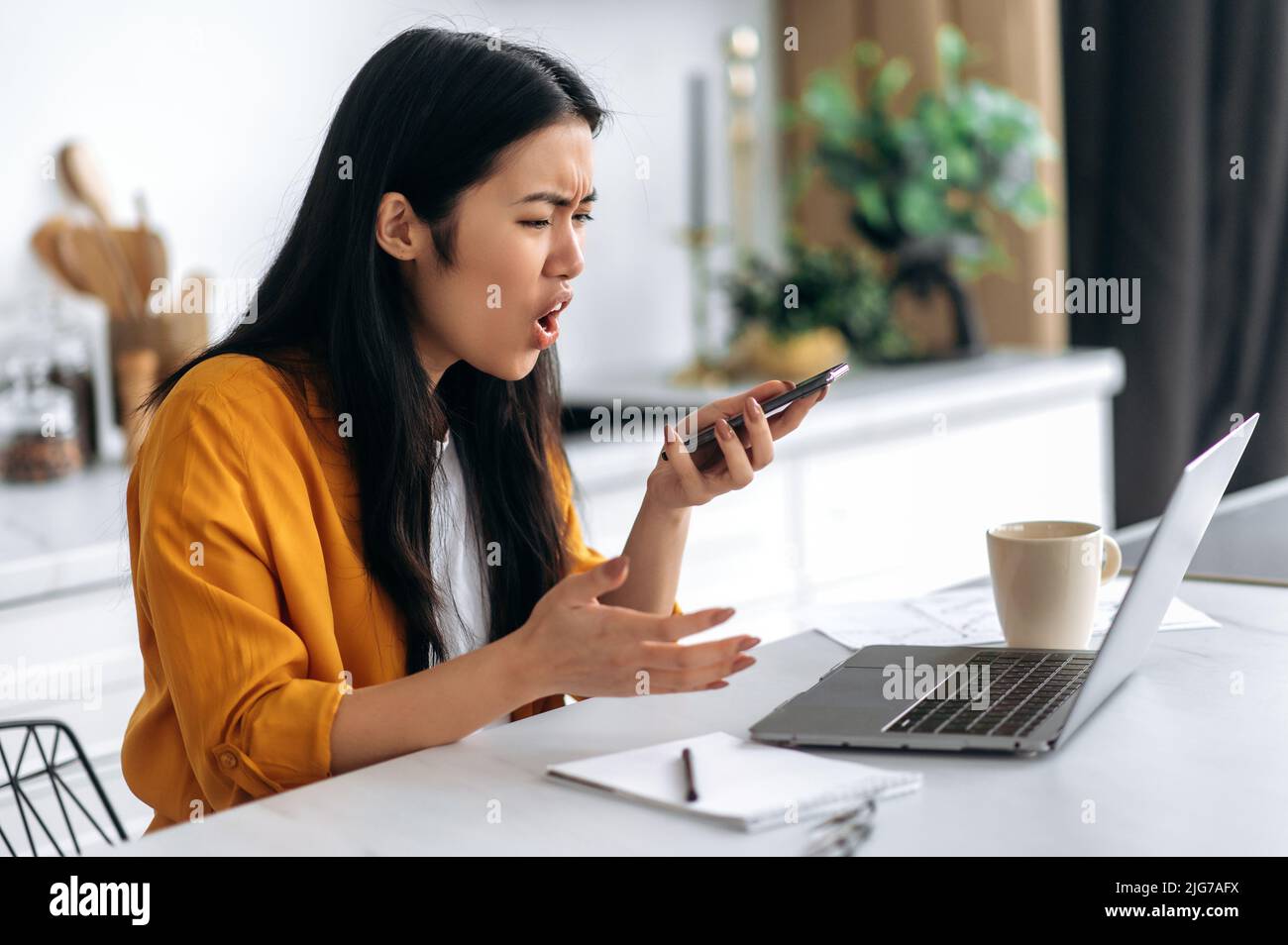 Infastidito giovane donna asiatica, freelance, studente, ceo, si siede a casa in cucina, lavoro a distanza, tenendo in mano lo smartphone parlare in vivavoce, registrare il messaggio audio, espressione emotiva del volto Foto Stock