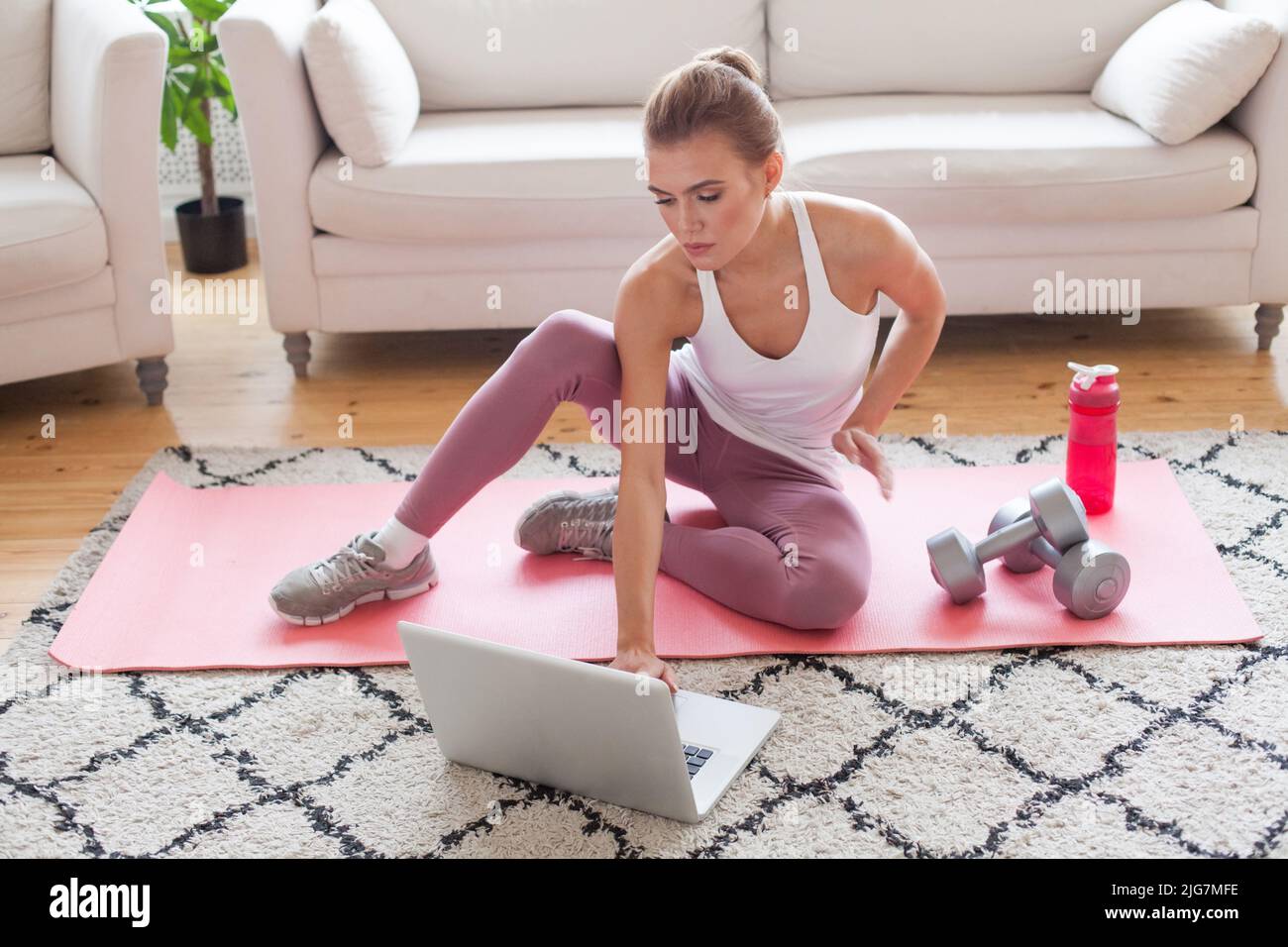 Donna allegra e sportiva seduta sul tappeto con manubri e bottiglia d'acqua e con un computer portatile a casa. Stile di vita sano. Soggiorno a casa attività Foto Stock