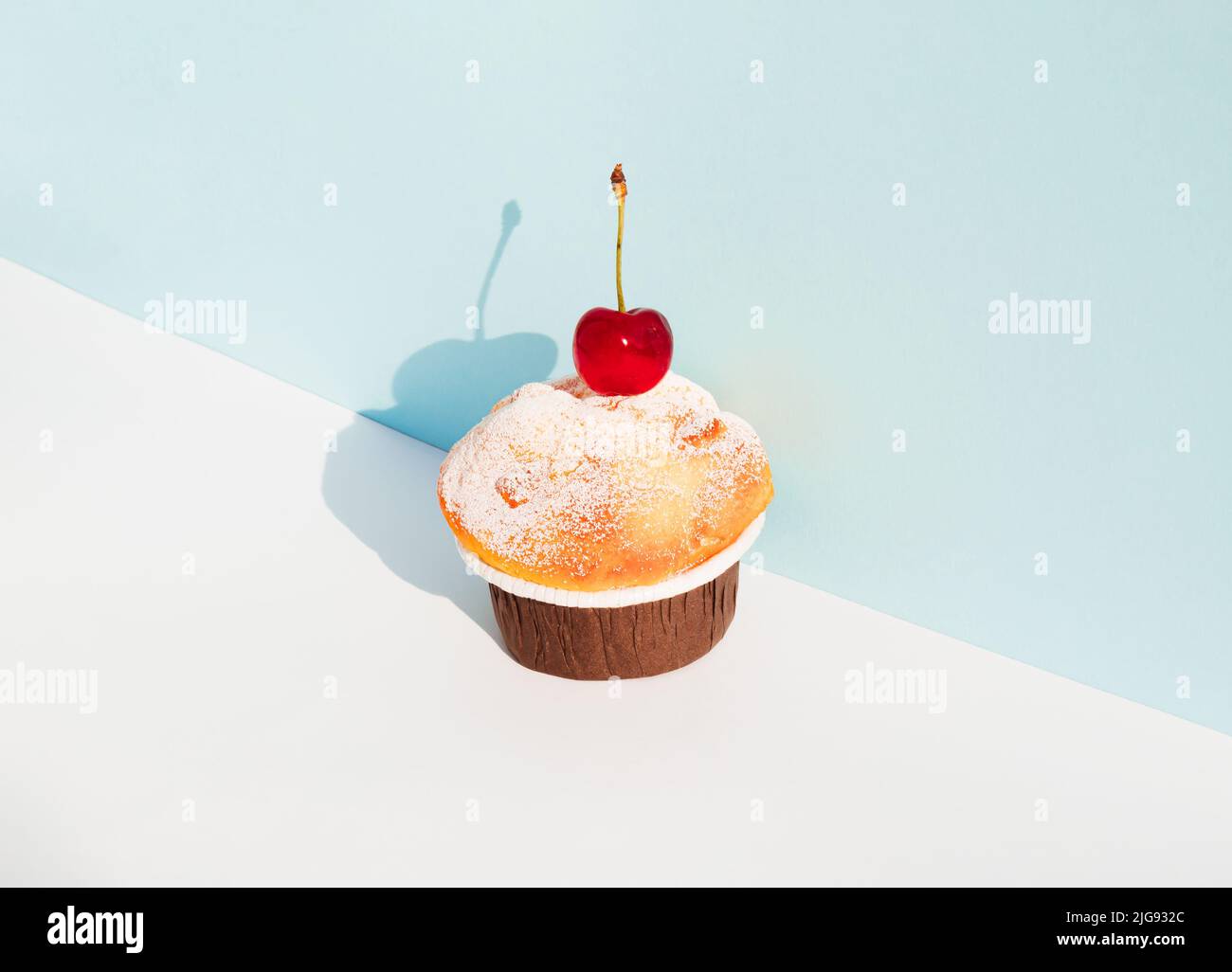Cupcake con ciliegia su sfondo bianco e blu pastello. Ombra di giorno soleggiata. Concetto minimo di cibo. Foto Stock Cupcake con ciliegia su sfondo bianco e blu pastello. Ombra di giorno soleggiata. Concetto minimo di cibo. Foto Stock