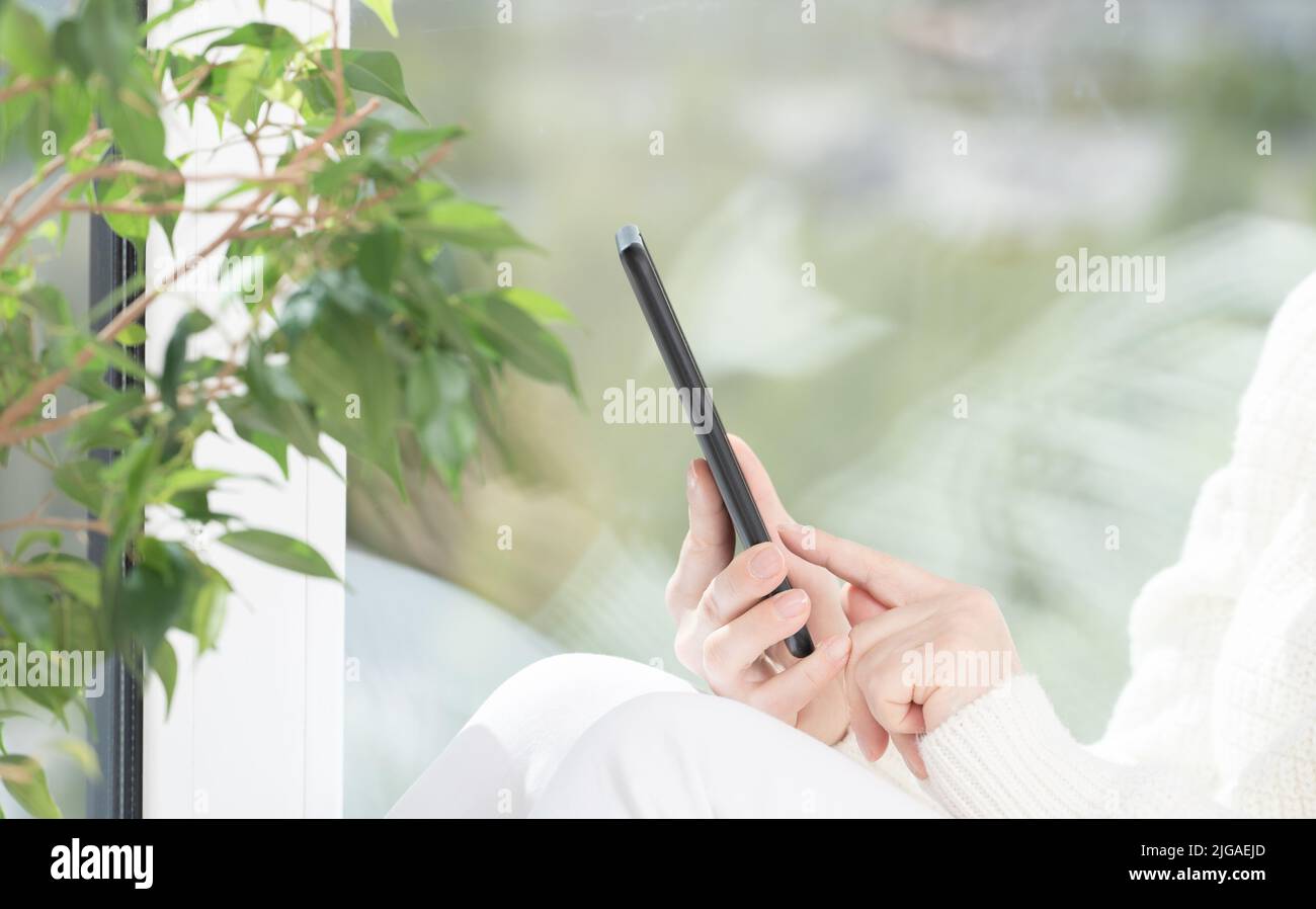 La ragazza si siede accanto alla finestra e usa il telefono cellulare, guarda lo smartphone, legge, acquista, comunica con gli amici in Messenger. Luce brillante immagine Foto Stock
