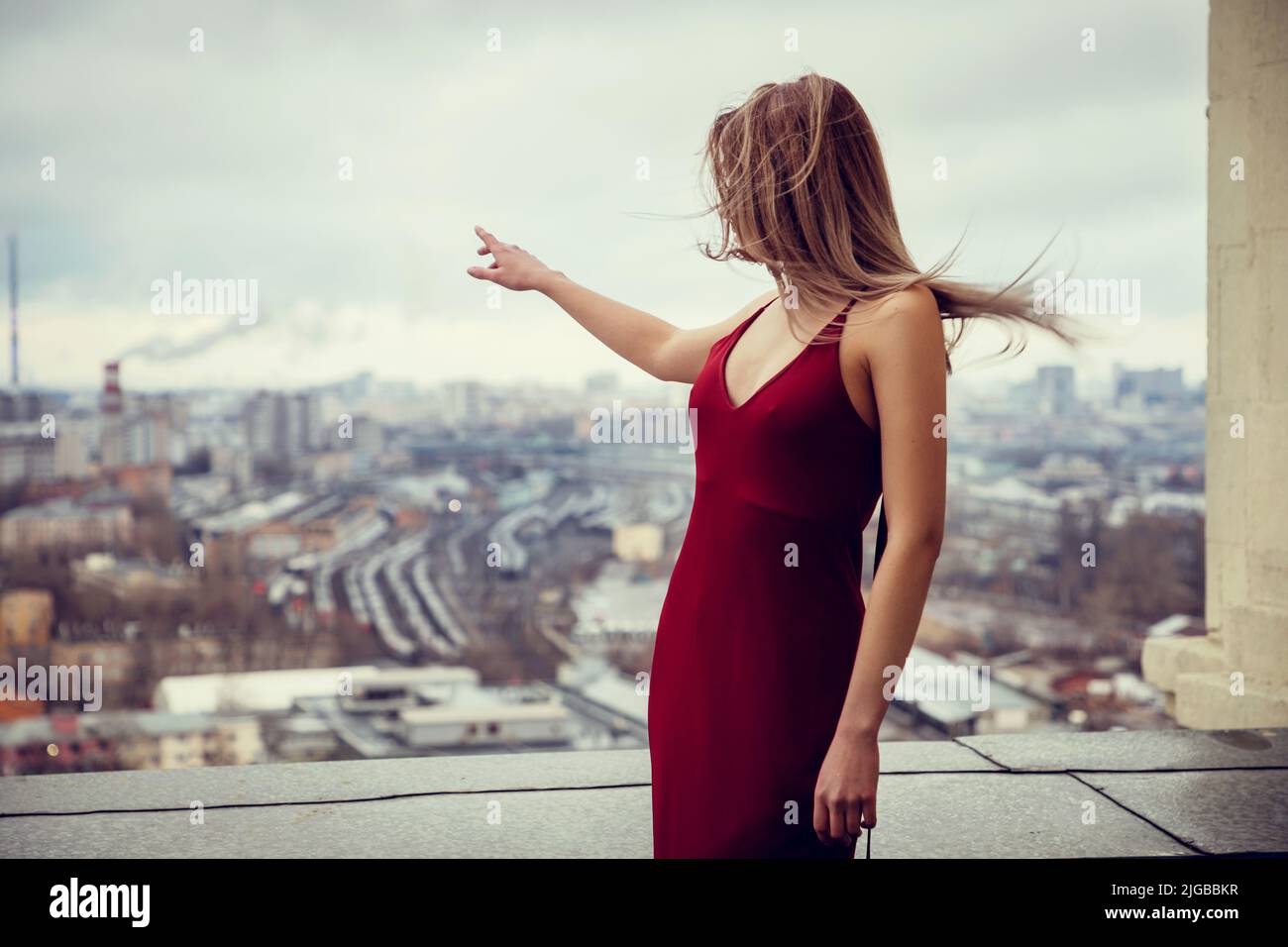 Ragazza sottile in abito rosso è in piedi sulla cima del grattacielo, alto sopra la megacità che si stende dietro di lei fino all'orizzonte. Punta con 1 mano Foto Stock