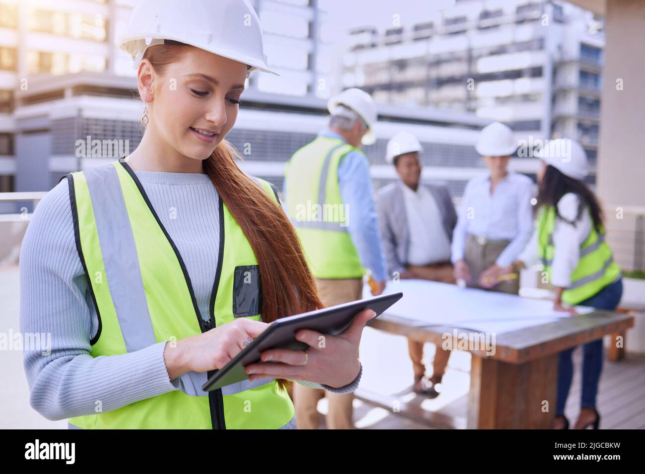 La costruzione è in corso. Una giovane e attraente lavoratrice di costruzioni che usa un tablet mentre si trova all'esterno con il suo team in background. Foto Stock