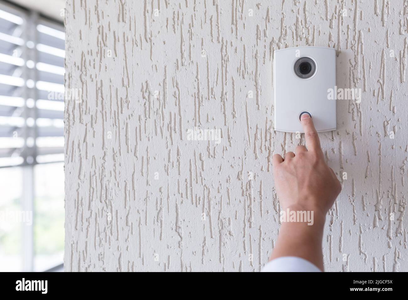 Mano di un uomo in una camicia bianca premendo il pulsante di una porta bussare su una parete bianca di una casa, un intelligente porta bussare con una videocamera per la comunicazione Foto Stock