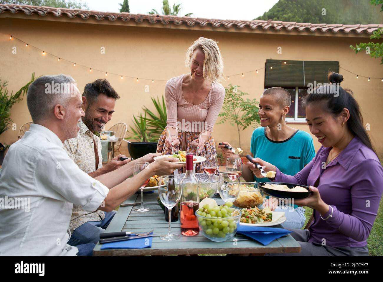 Donna caucasica di mezza età piuttosto sorridente che mette un piatto con cibo sul tavolo da pranzo in una festa con amici felici nel cortile. Foto Stock