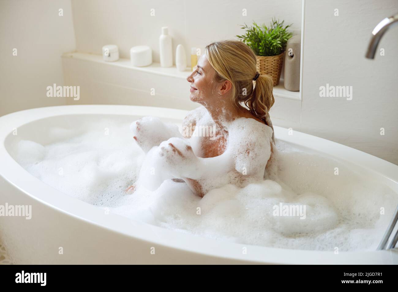 Gioiosa giovane attraente donna seduta in bagno di schiuma e bolle e sorridente. Fare il bagno. Foto Stock