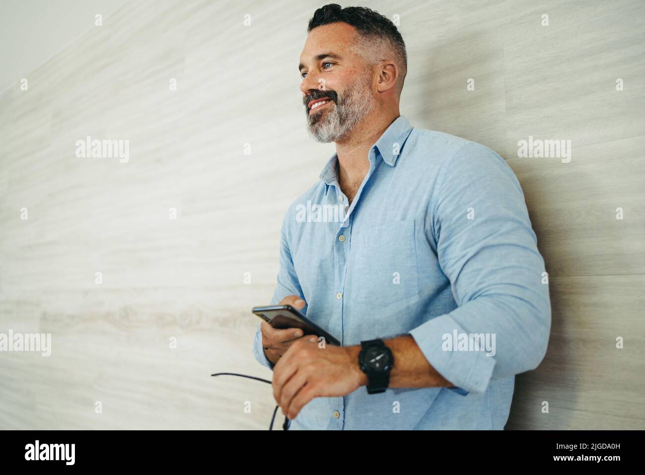 Un uomo d'affari felice e maturo sorridente e che guarda via mentre si trova in un ufficio moderno. Uomo d'affari maturo e premuroso che tiene uno smartphone in piedi Foto Stock
