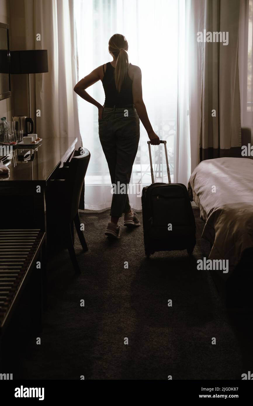 Donna che viaggia con valigia dopo il check-in in una camera d'hotel che guarda alla finestra. Silhouette di visitatore in alloggio di viaggio pronto Foto Stock
