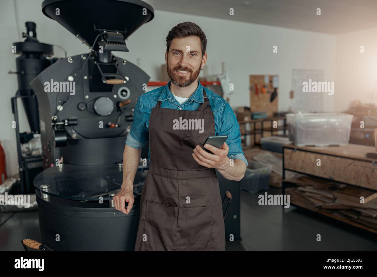 Barista sorridente con telefono in piedi sullo sfondo della macchina per la tostatura del caffè Foto Stock