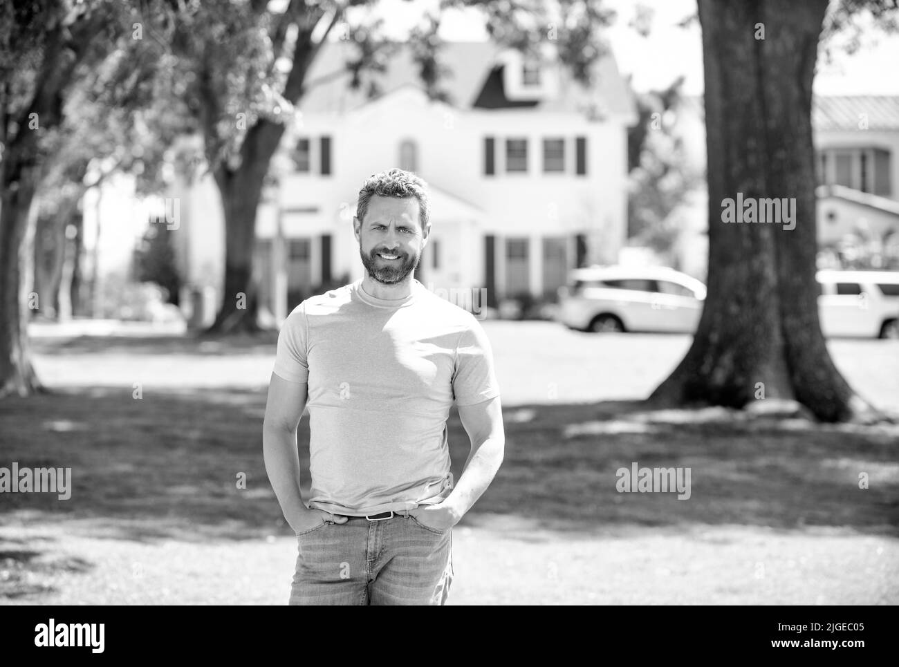 felice uomo barbuto in piedi su sfondo casa, proprietà Foto Stock