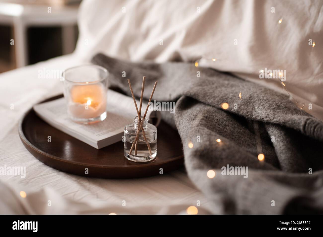 Home fragranza liquida su libro di carta aperto e candela profumata bruciante su vassoio di legno e maglione di stoffa a maglia in letto da vicino. Atmosfera accogliente. Foto Stock