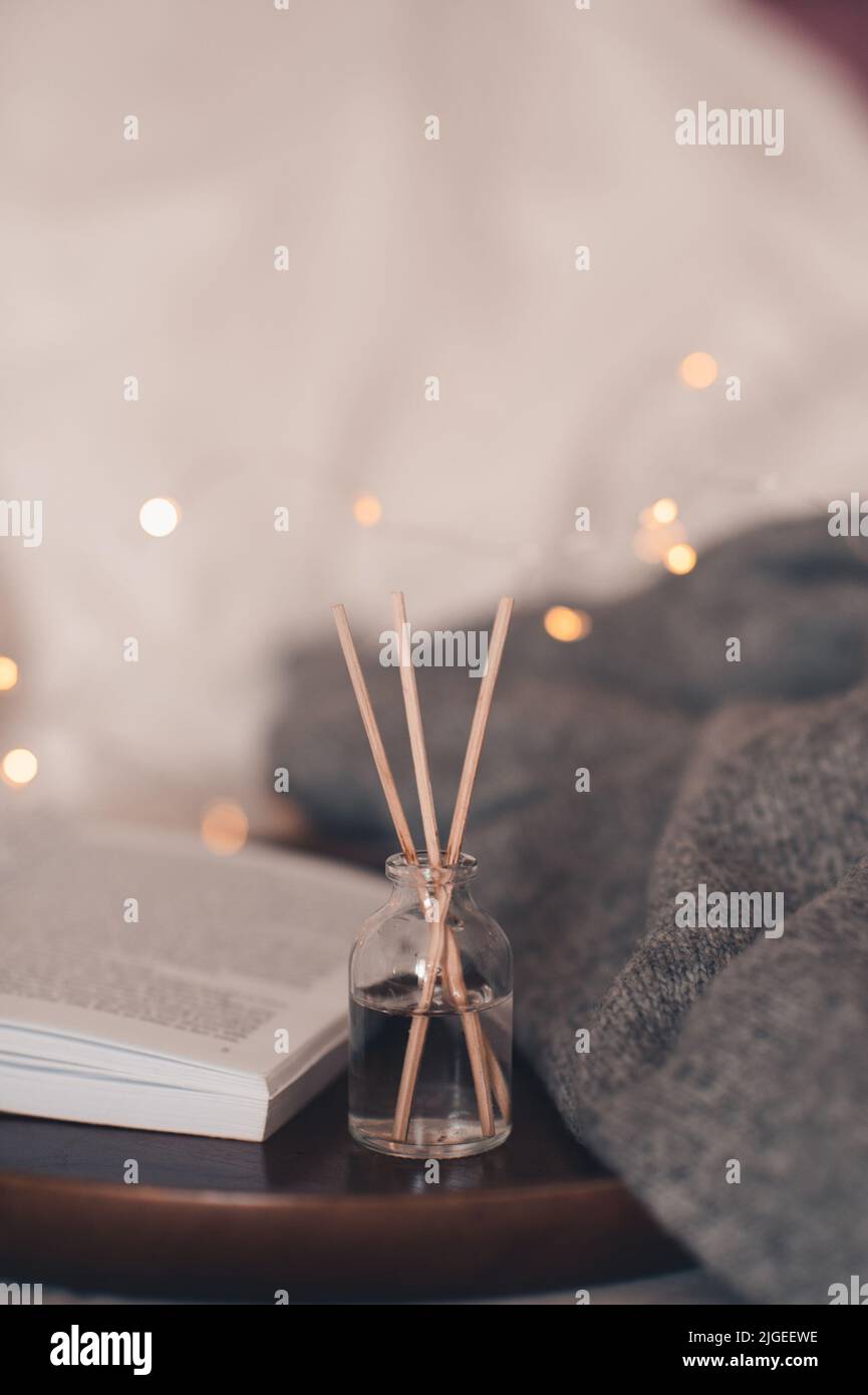 Home fragranza liquida su libro di carta aperto e candela profumata bruciante su vassoio di legno e maglione di stoffa a maglia in letto da vicino. Atmosfera accogliente. Foto Stock