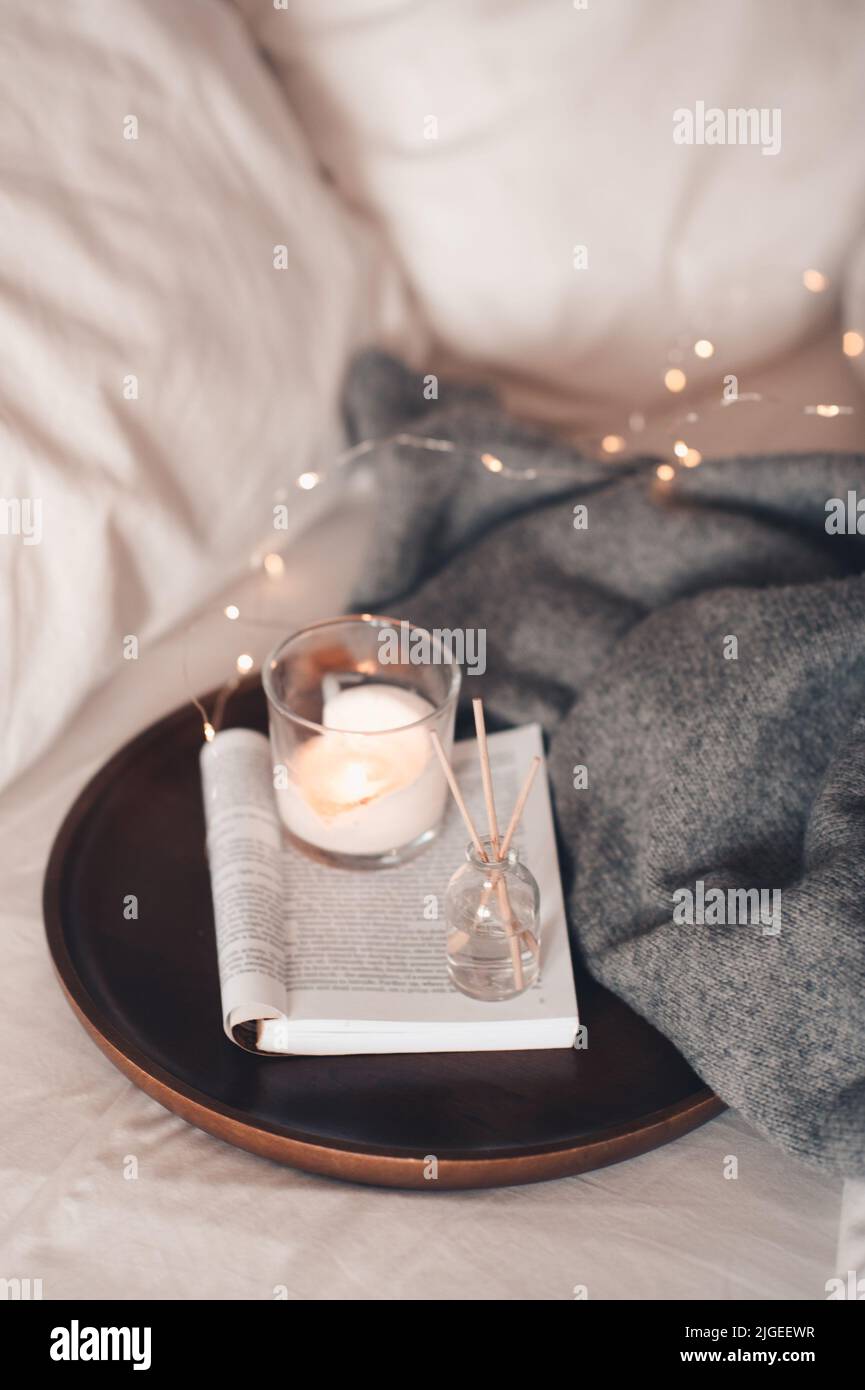 Home fragranza liquida su libro di carta aperto e candela profumata bruciante su vassoio di legno e maglione di stoffa a maglia in letto da vicino. Atmosfera accogliente. Foto Stock