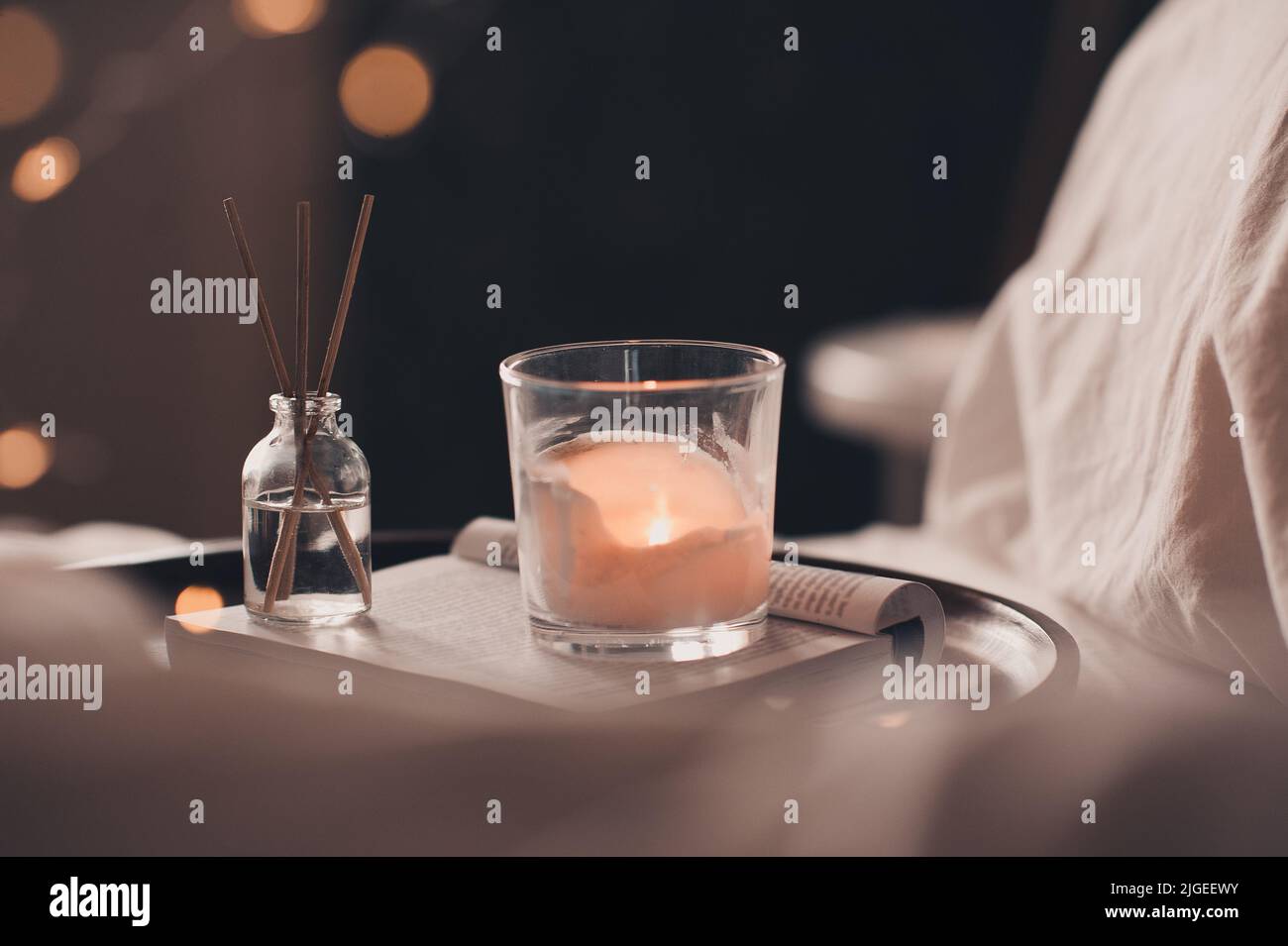 Home fragranza liquida su libro di carta aperto e candela profumata bruciante su vassoio di legno e maglione di stoffa a maglia in letto da vicino. Atmosfera accogliente. Foto Stock