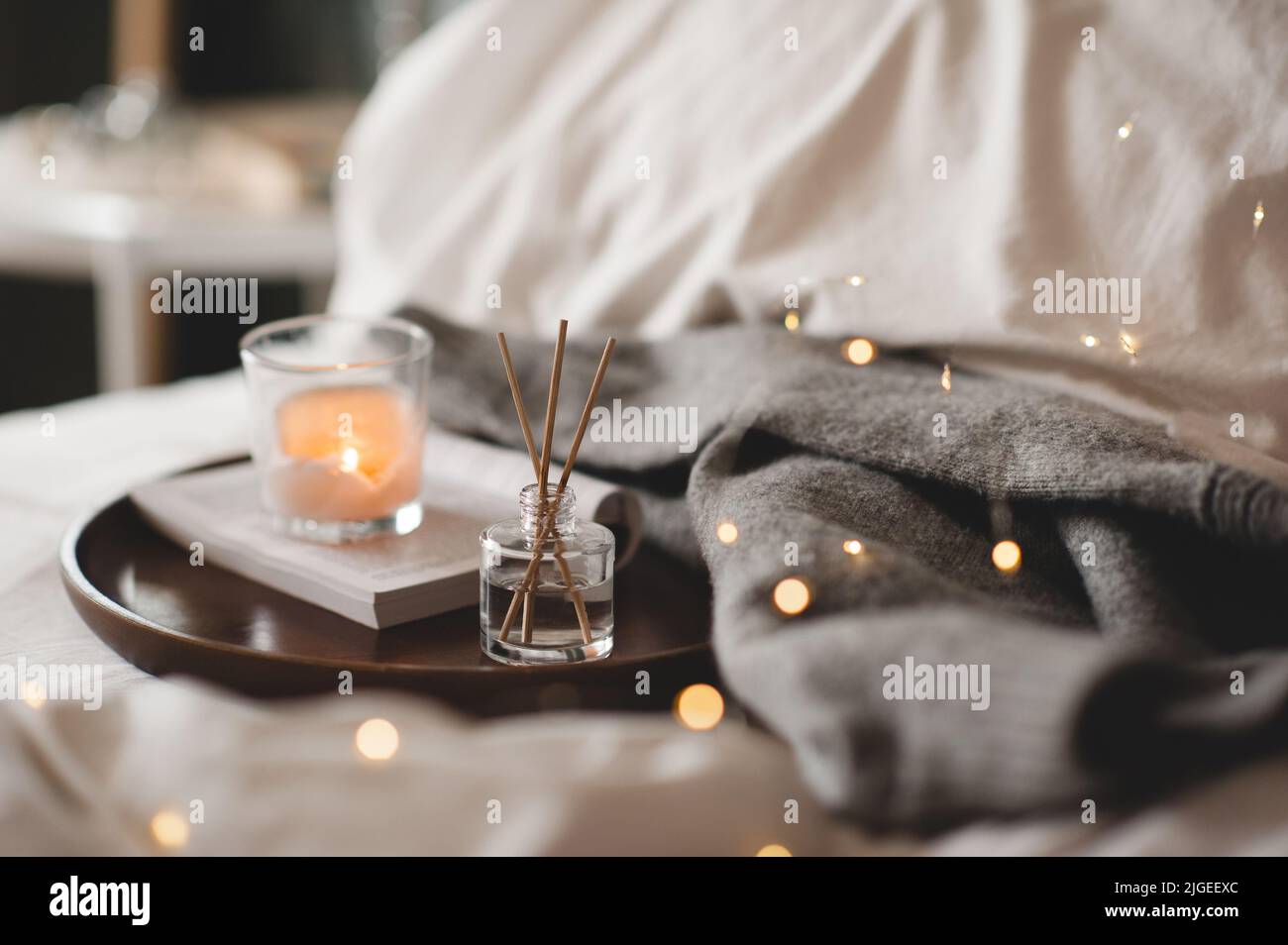 Home fragranza liquida su libro di carta aperto e candela profumata bruciante su vassoio di legno e maglione di stoffa a maglia in letto da vicino. Atmosfera accogliente. Foto Stock