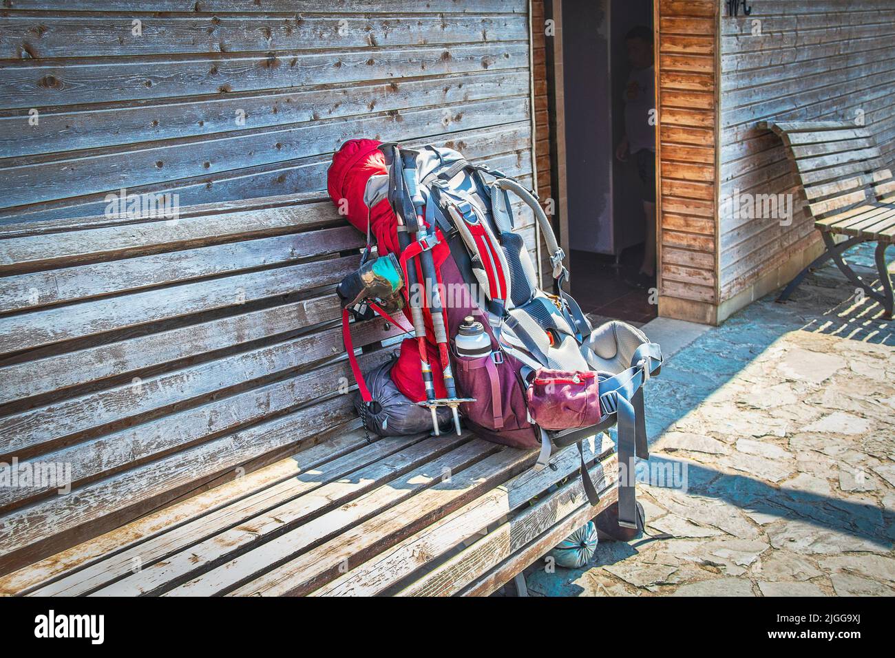 Grande zaino carico con bastoni da passeggio e bottiglia d'acqua e sacco a pelo e seggiolino per bambini su panca di legno al di fuori dei bagni in legno - escursioni Foto Stock