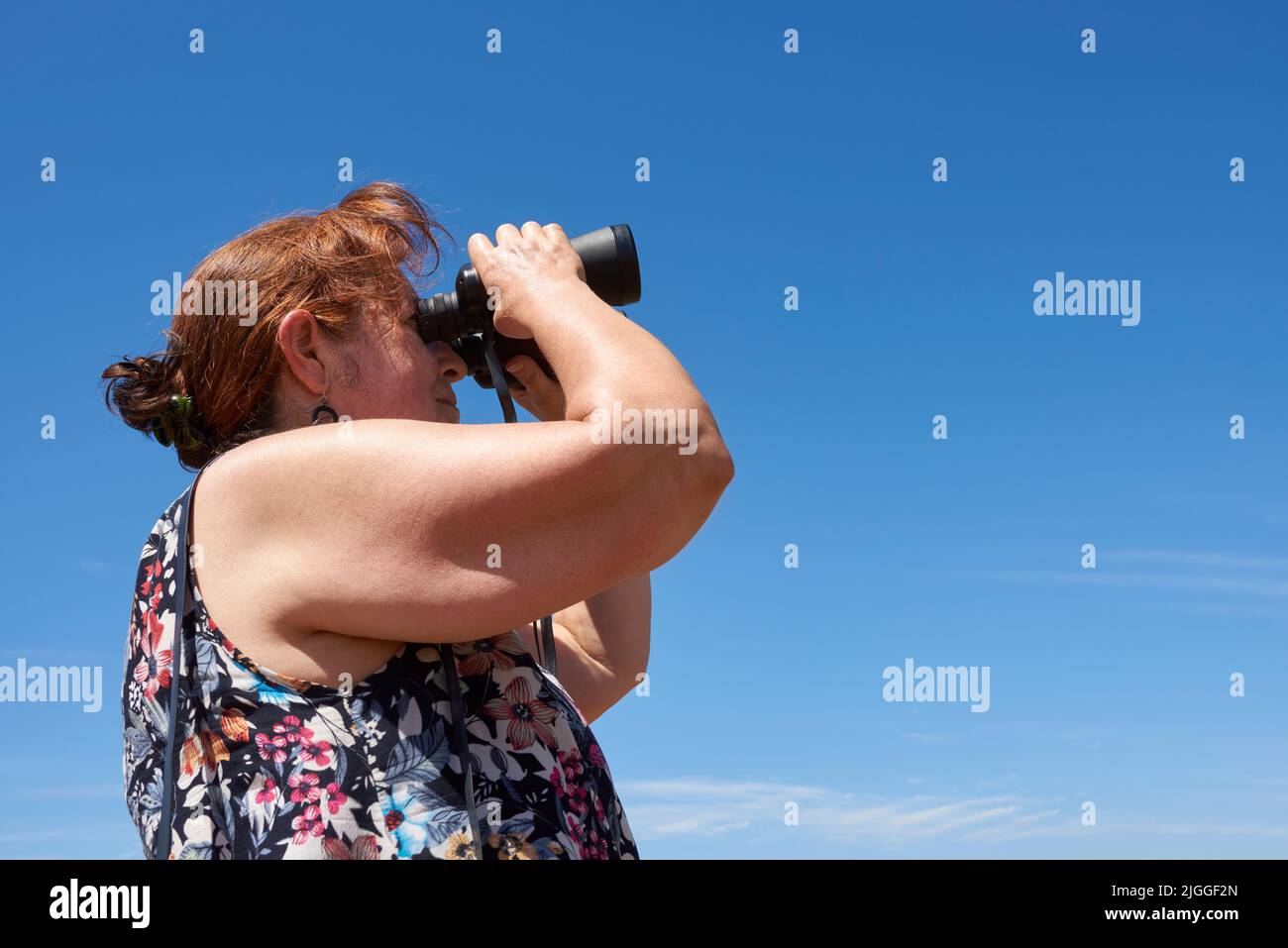 Donna ispanica matura guardando in lontananza utilizzando binocoli all'aperto in una giornata estiva. Concetti: Avventura, viaggio nella natura, guardare nel futuro Foto Stock