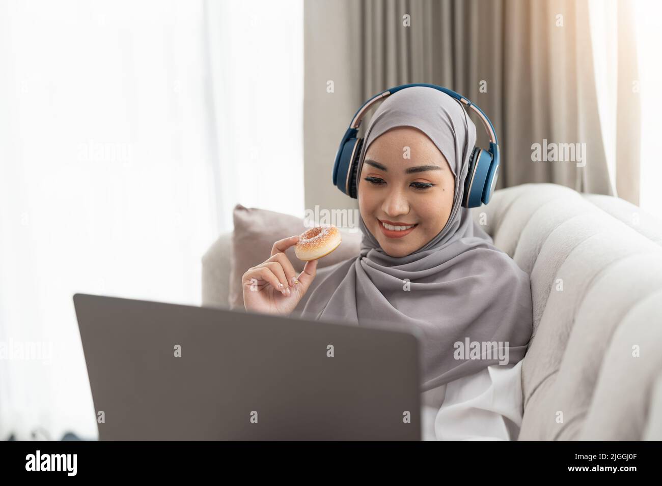 Una bella e rilassata donna musulmana asiatica che indossa un hijab e cuffie sta guardando un film sul suo computer portatile mentre sorseggiando tè caldo in vita Foto Stock