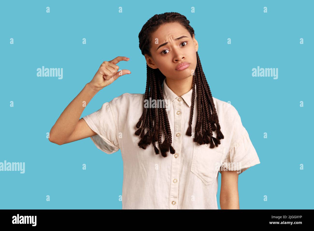 Donna infelice attraente con dreadlock neri che mostrano piccole dimensioni, in piedi dimostra piccola cosa o oggetto, indossando camicia bianca. Studio interno girato isolato su sfondo blu. Foto Stock