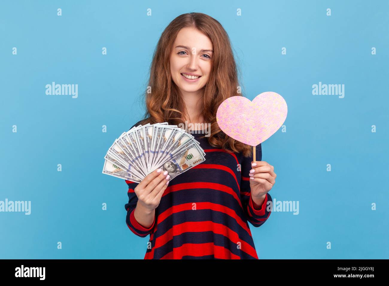 Donna felice indossando a righe in stile casual maglione, in piedi grande fan di banconote in dollari e cuore rosa sul bastone, esprimendo l'amore per i soldi, Indoor studio girato isolato su sfondo blu. Foto Stock