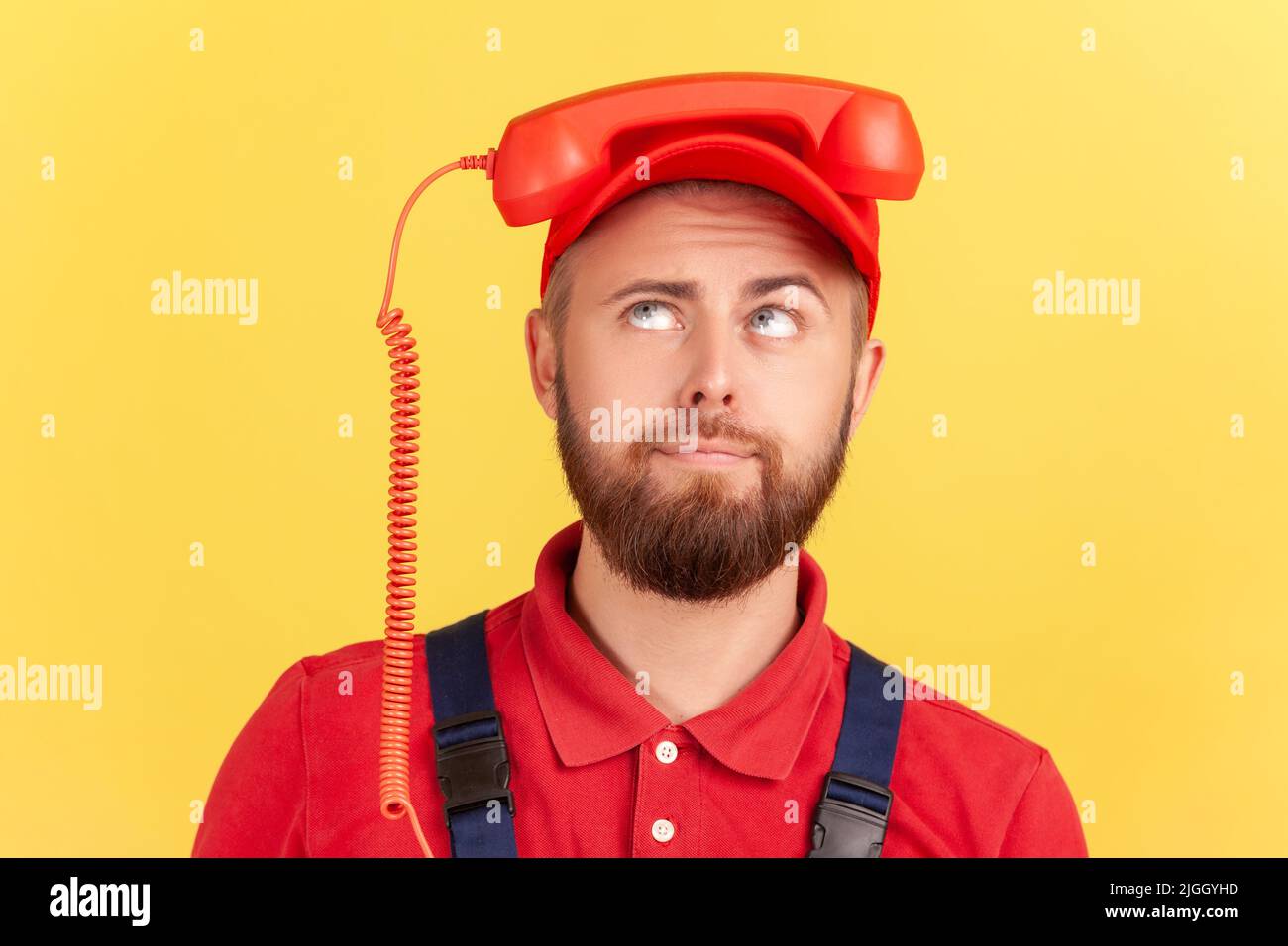 Ritratto di uomo operaio divertente che tiene la cornetta sulla sua testa e guardando in su, stanco di chiamare servizio di ordine, indossare tute e cappuccio rosso. Studio interno girato isolato su sfondo giallo. Foto Stock