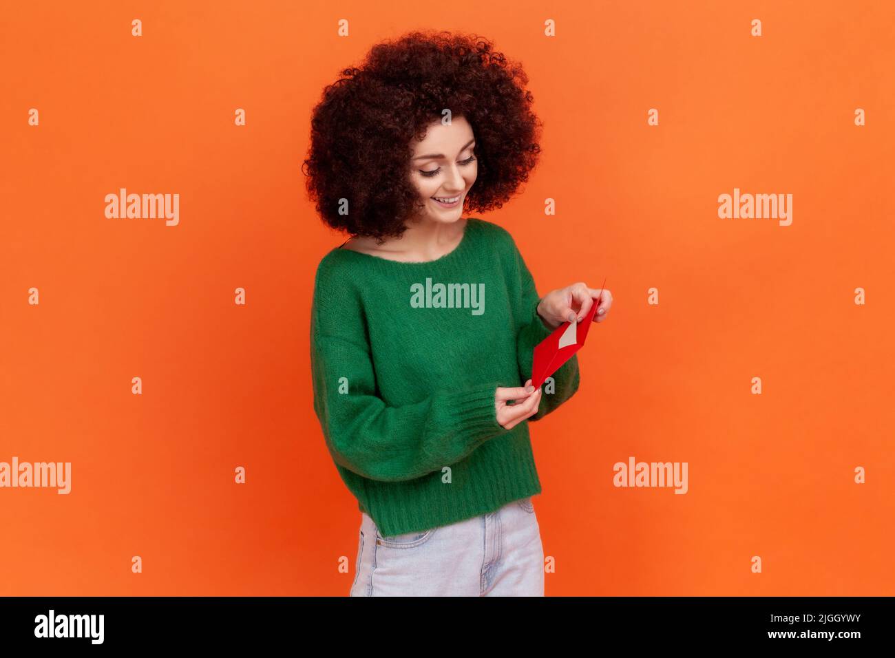 Ritratto di donna positiva con acconciatura Afro indossando il maglione verde casual stile in piedi tenendo la busta rossa, lettura lettera romantica. Studio interno girato isolato su sfondo arancione. Foto Stock