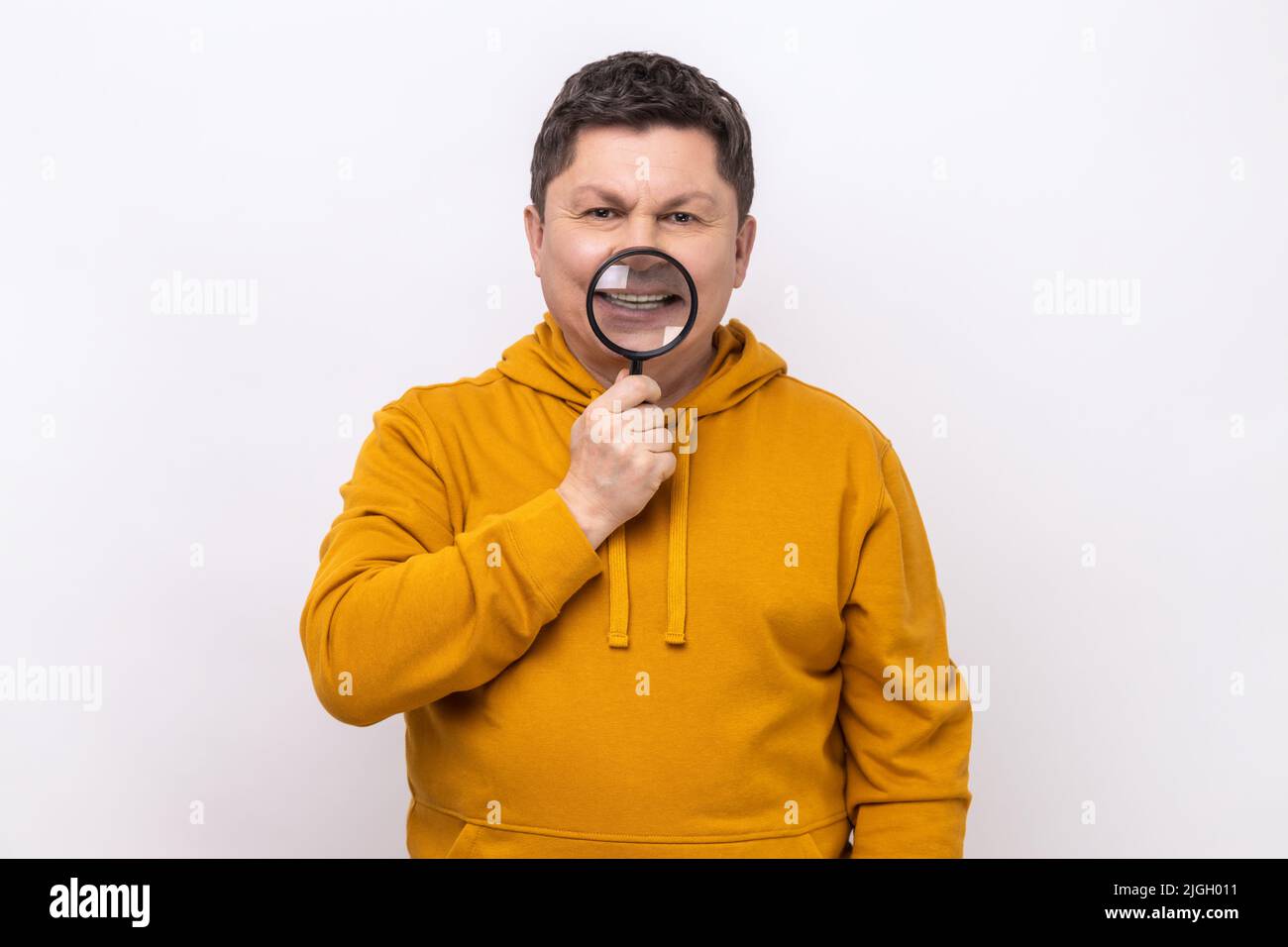 Ritratto di uomo di mezza età in piedi, tenendo la lente d'ingrandimento sui denti, guardando con un sorriso toothy, indossando una felpa con cappuccio urban style. Studio interno girato isolato su sfondo bianco. Foto Stock