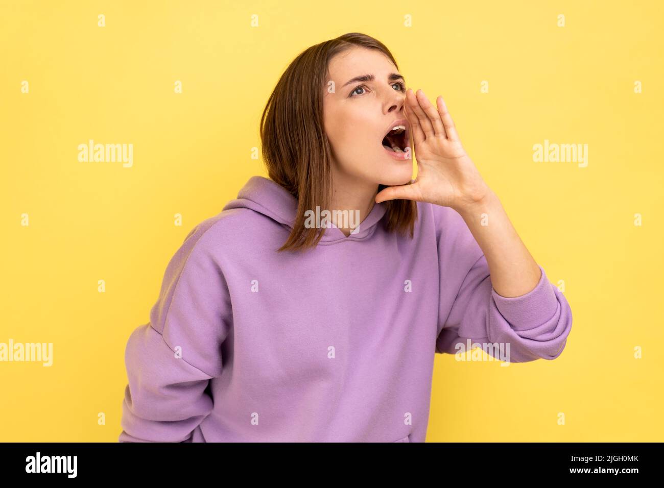 Attenzione, pubblicità. Profilo di donna urlando gridando circa la vendita, annunciando ad alta voce messaggio importante, indossando felpa con cappuccio viola. Studio interno girato isolato su sfondo giallo. Foto Stock