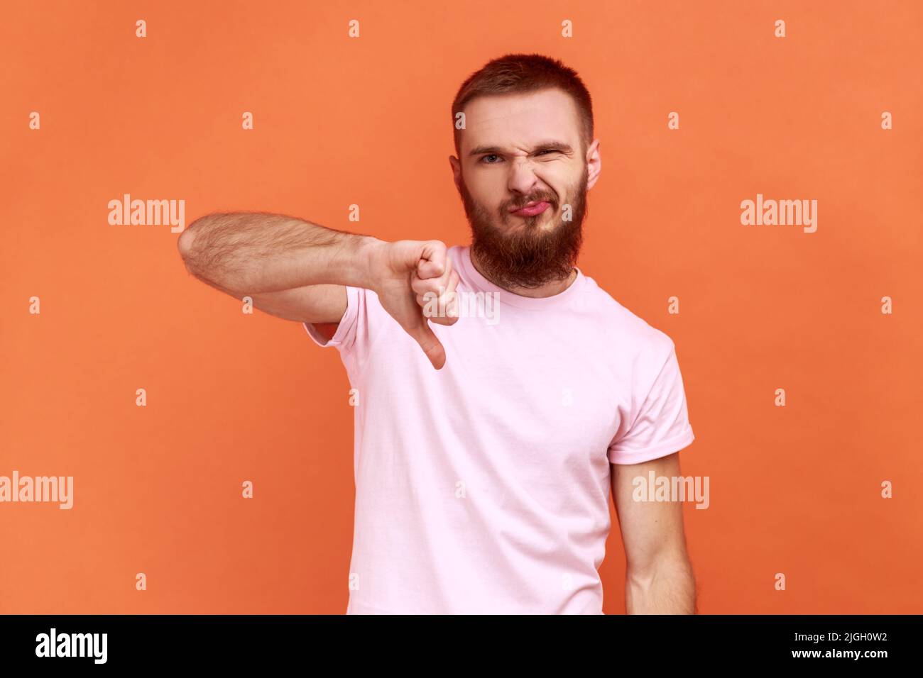 Ritratto di uomo criticando la cattiva qualità con i pollici giù soppiantò la grimace, mostrando gesto dissimile, esprimendo disapprovazione, indossando la T-shirt rosa. Studio interno girato isolato su sfondo arancione. Foto Stock
