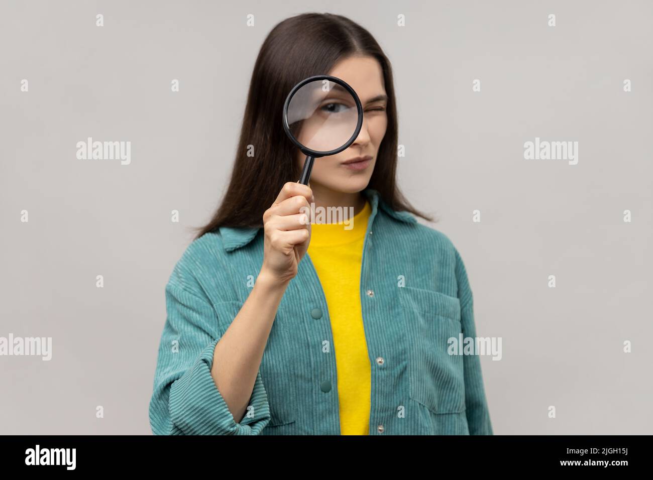 Donna in piedi, con lente d'ingrandimento e fotocamera con grande zoom, espressione concentrata, indossando una giacca casual. Studio interno girato isolato su sfondo grigio. Foto Stock