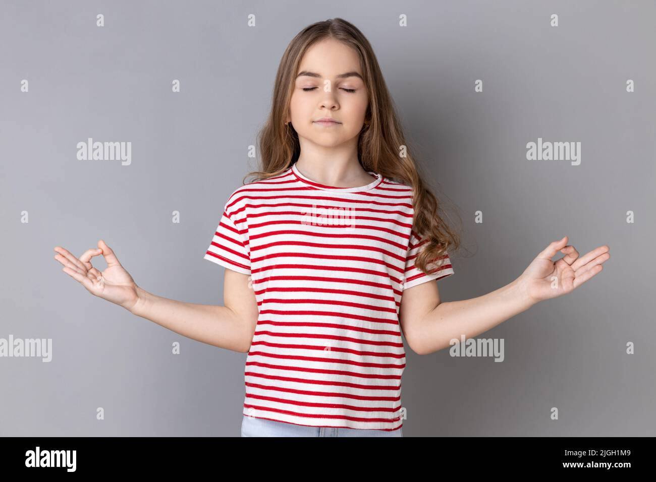 Ritratto di calma rilassata ragazza con t-shirt a righe che fa yoga, respirando, mantenendo gli occhi chiusi, calmando dopo la scuola. Studio interno girato isolato su sfondo grigio. Foto Stock