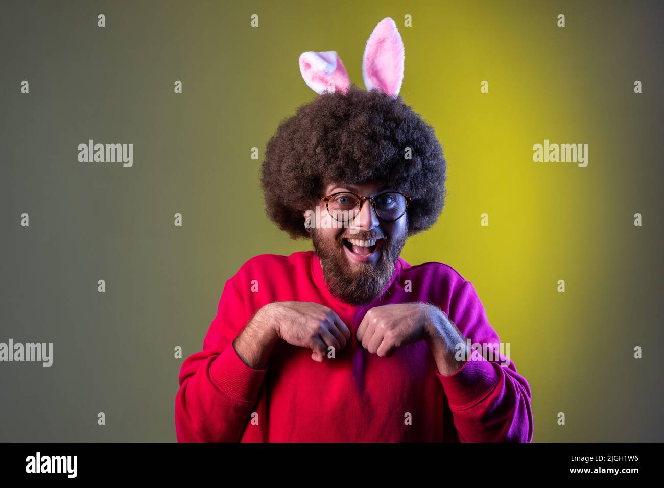 Stupito uomo di hipster eccitato con acconciatura afro e orecchie conigliate, facendo zampe con le mani, guardando la macchina fotografica, indossando una felpa rossa. Studio interno girato isolato su colorato sfondo di luce al neon. Foto Stock